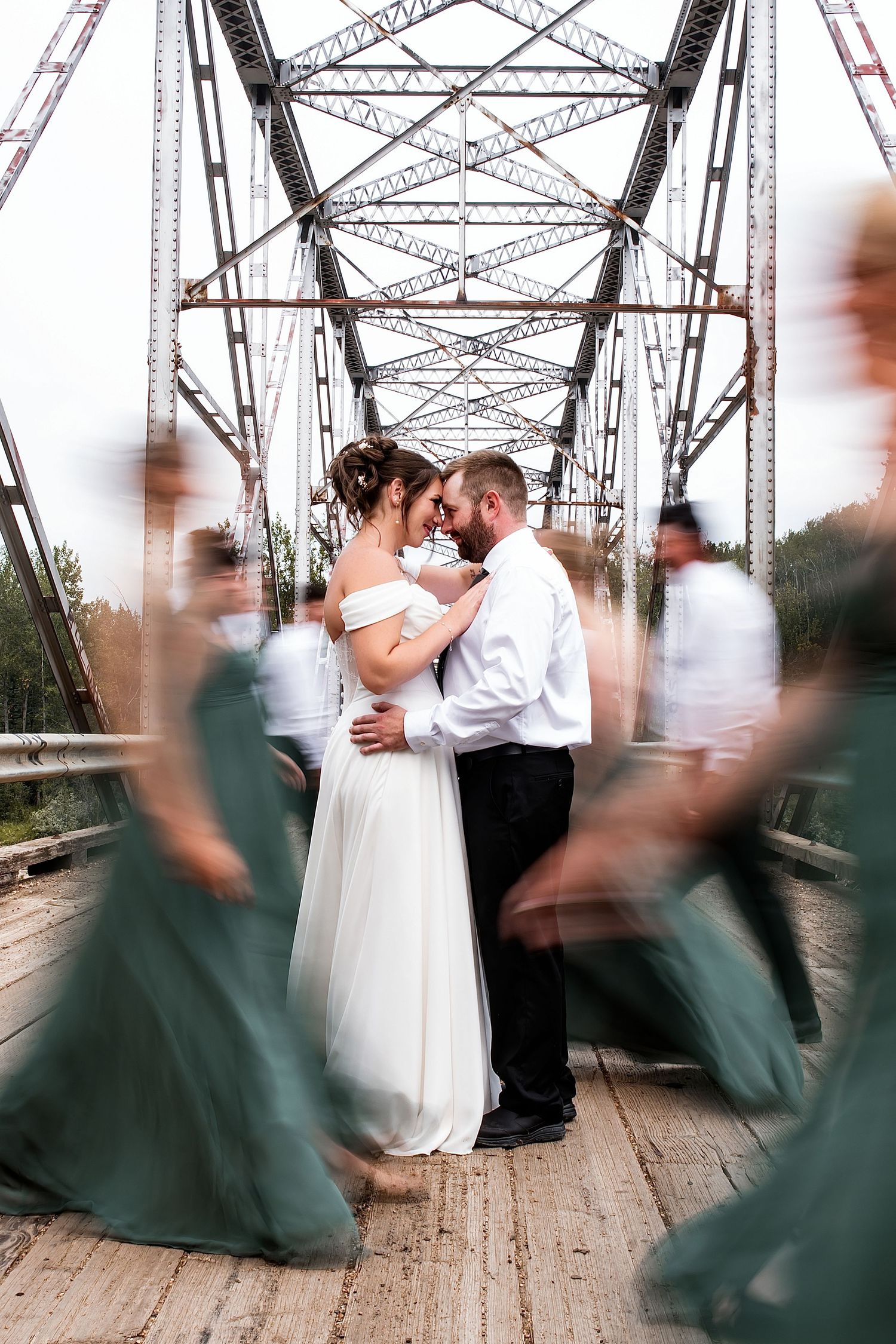 Bride and groom share intimate moment on rustic iron bridge, surrounded by blurred bridesmaids in green gowns.