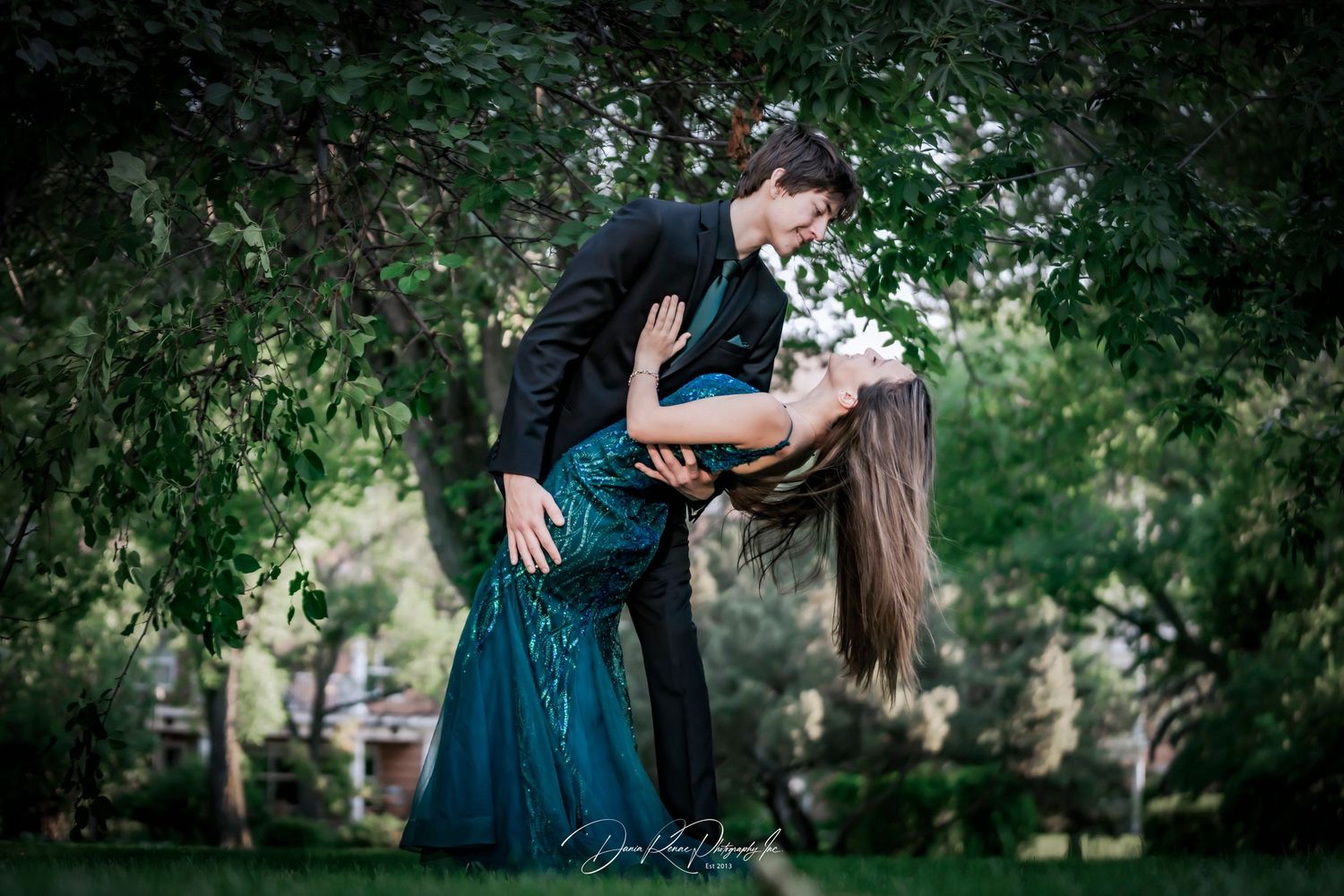 A couple in formal attire shares a romantic dip kiss outdoors among lush green trees at prom.