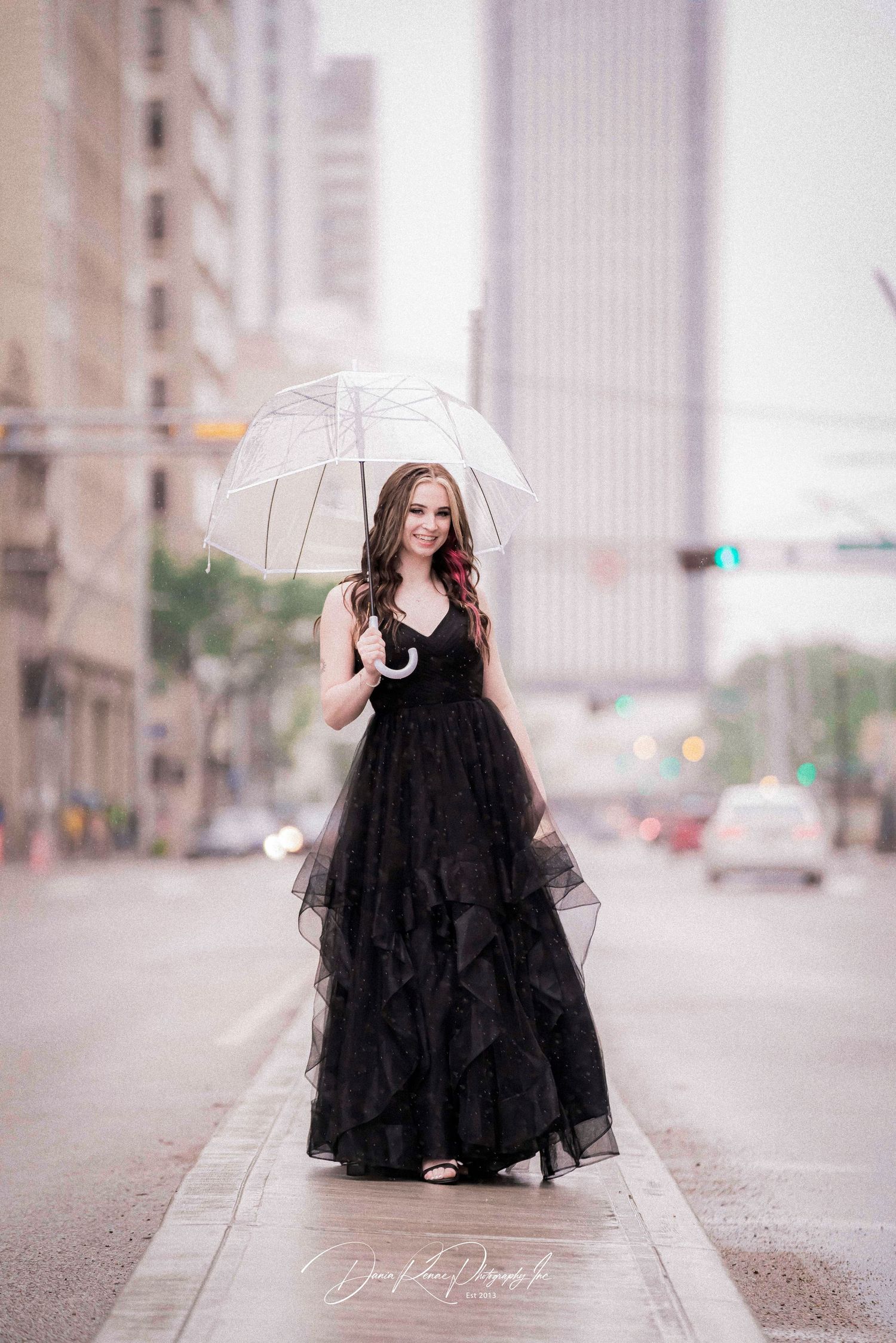 A woman in a black ruffled gown holds a clear umbrella on a rainy city street, posing elegantly.