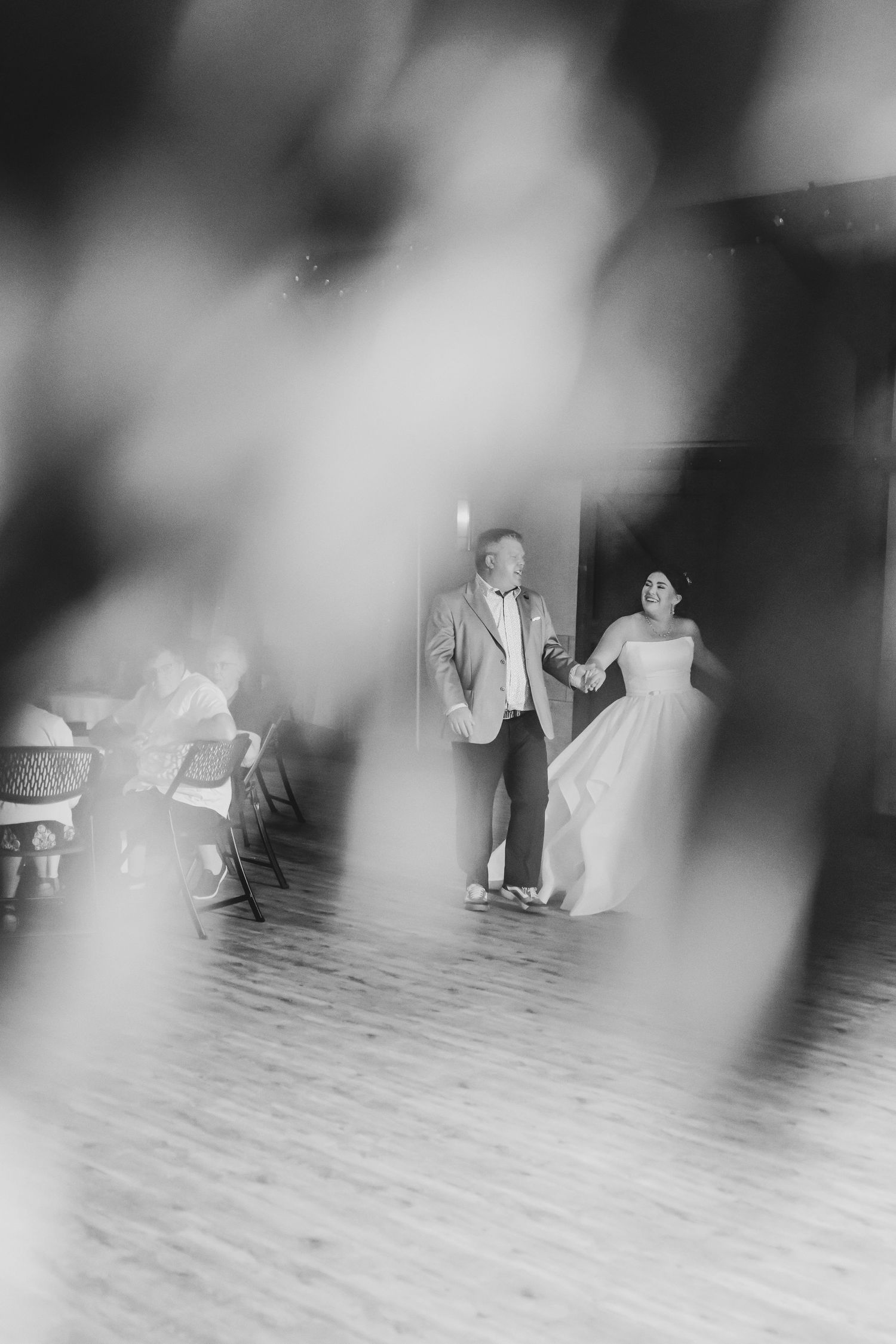 Black and white photo of bride and groom holding hands, walking onto wedding reception dance floor.