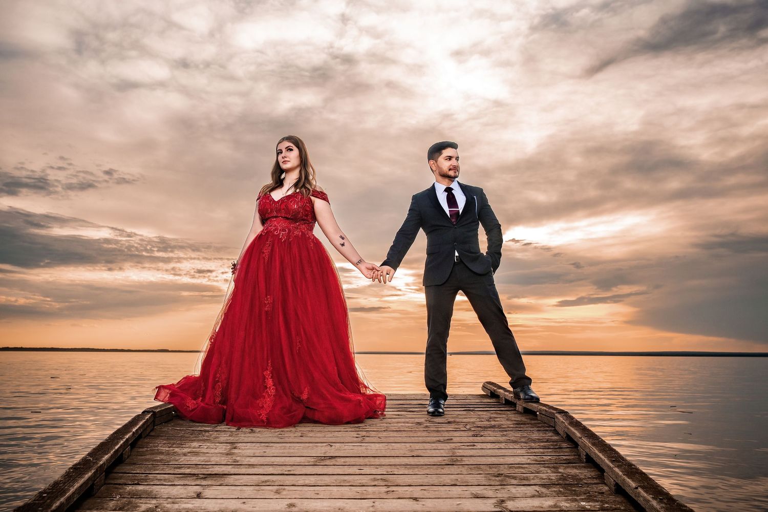 Couple in formal wear holding hands on a wooden pier at sunset, woman in red gown, man in black suit.