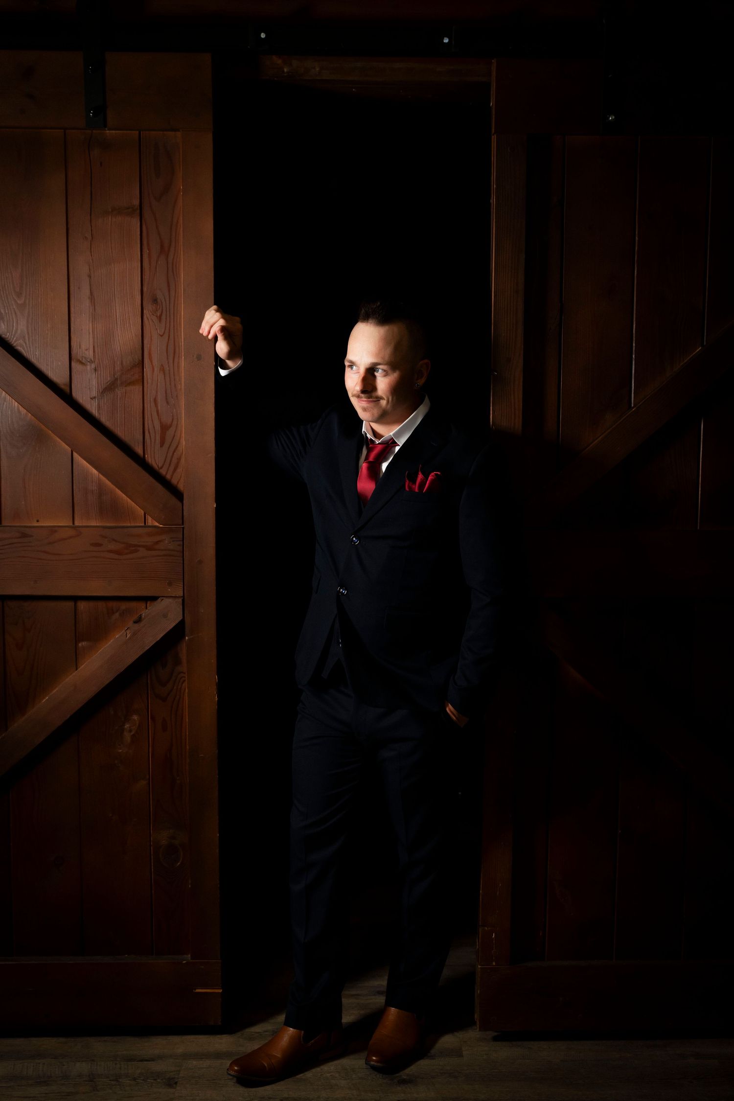 Man in navy suit with red tie and pocket square stands confidently in rustic barn doorway.