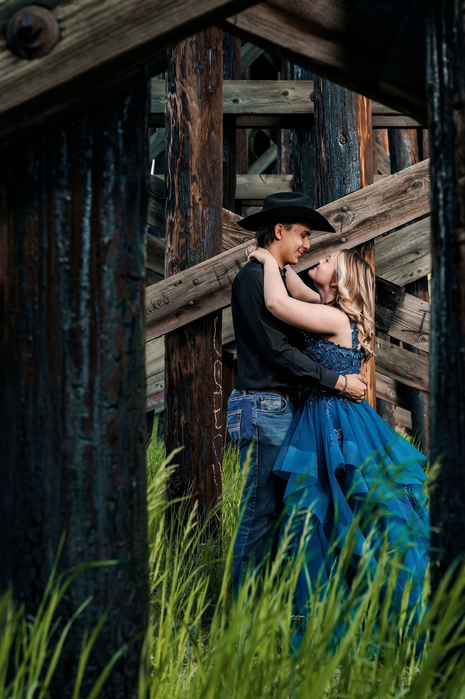 Couple embracing romantically near rustic wooden barn, man in cowboy hat and woman in blue dress, surrounded by tall green grass.
