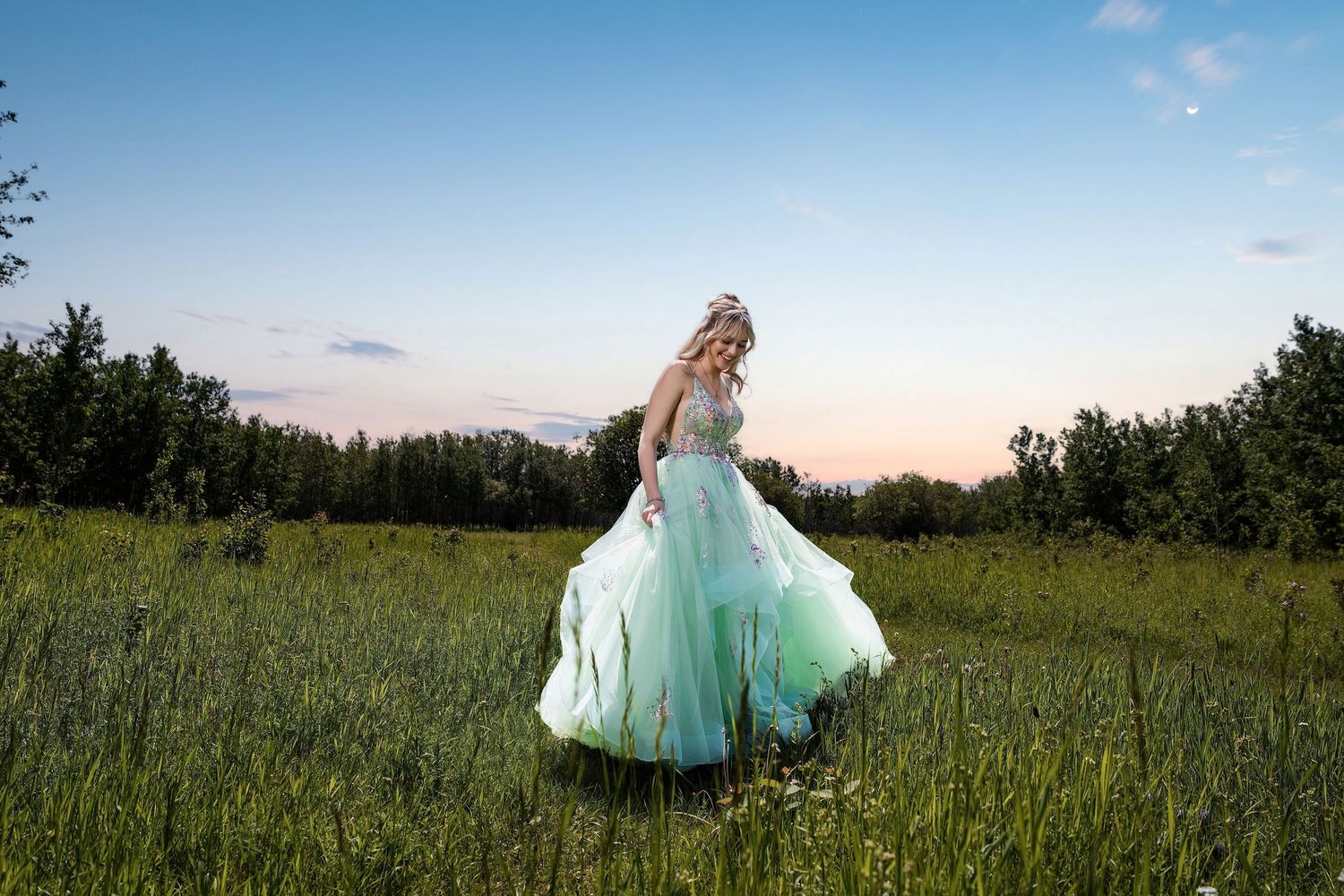 A woman in a flowing mint green ball gown twirls in a lush meadow at dusk with trees in the background.