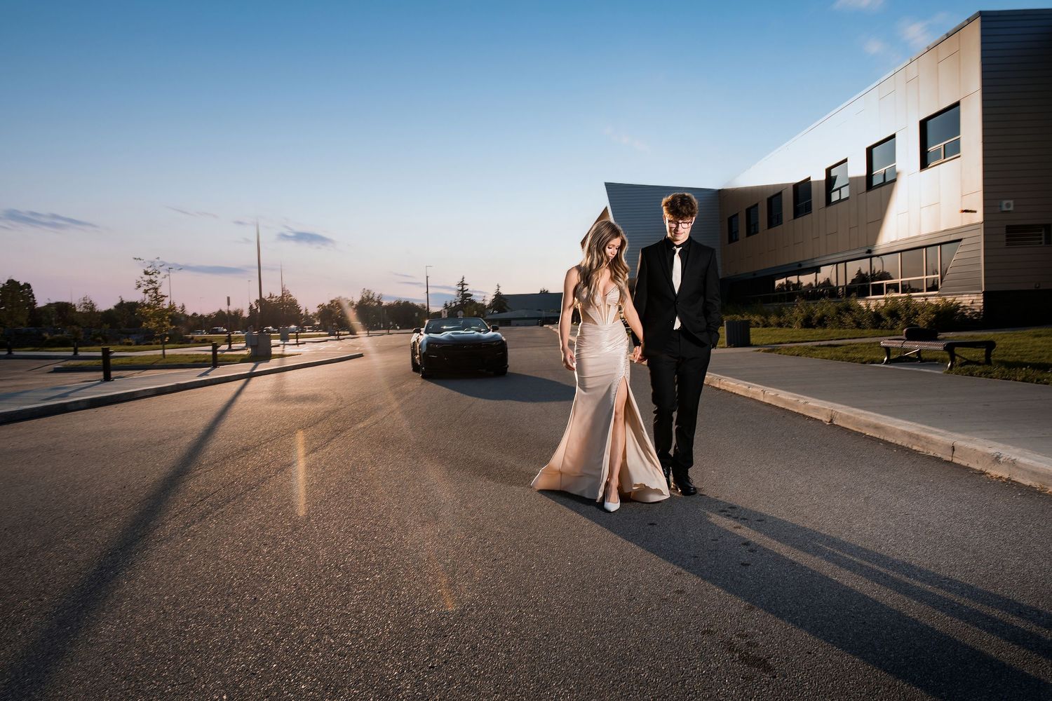 Elegant couple in formal attire walking on a road at sunset, with a luxury car and modern building in the background.