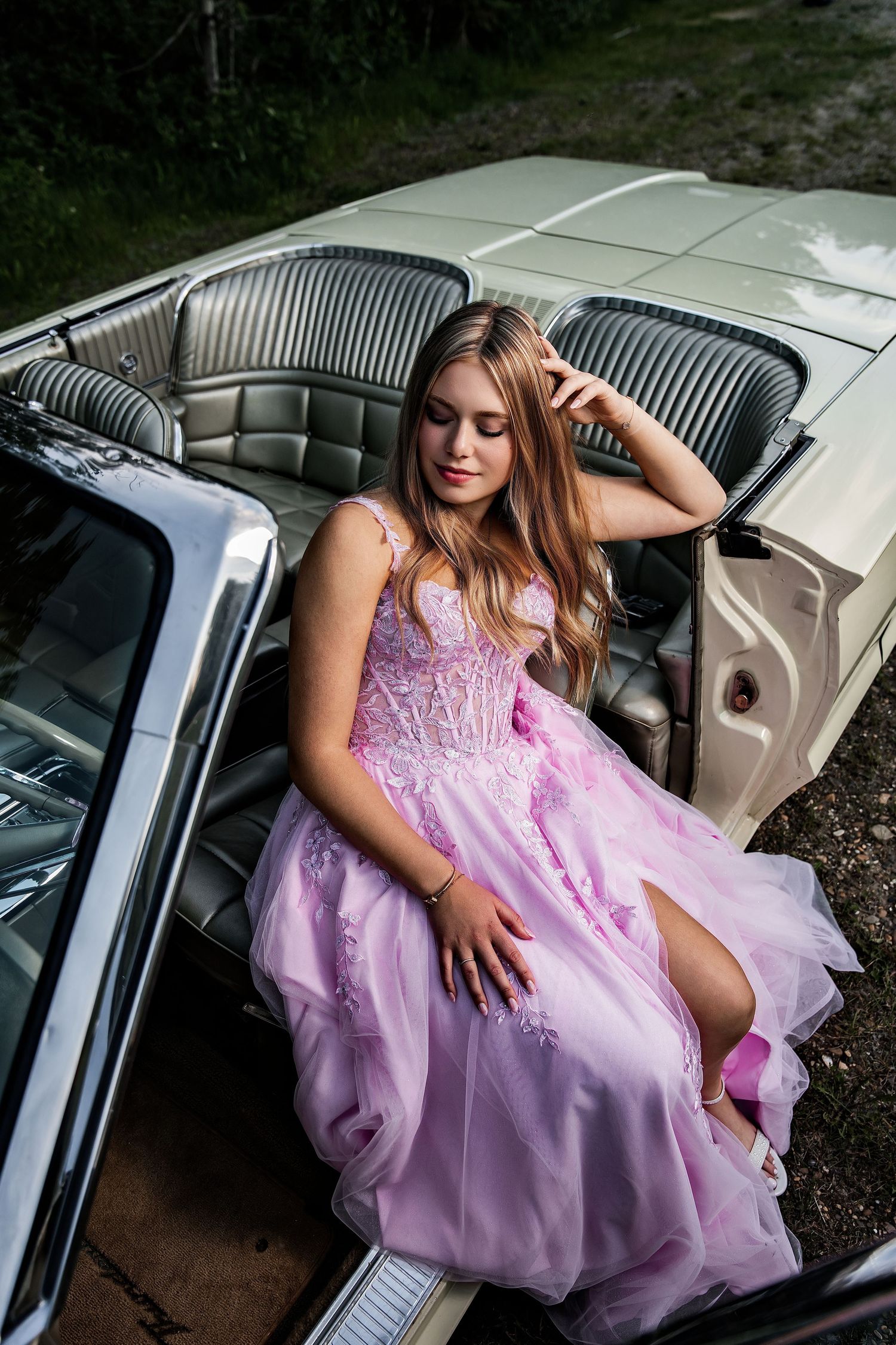 Young woman in pink ball gown posing in vintage white convertible car for prom photos.