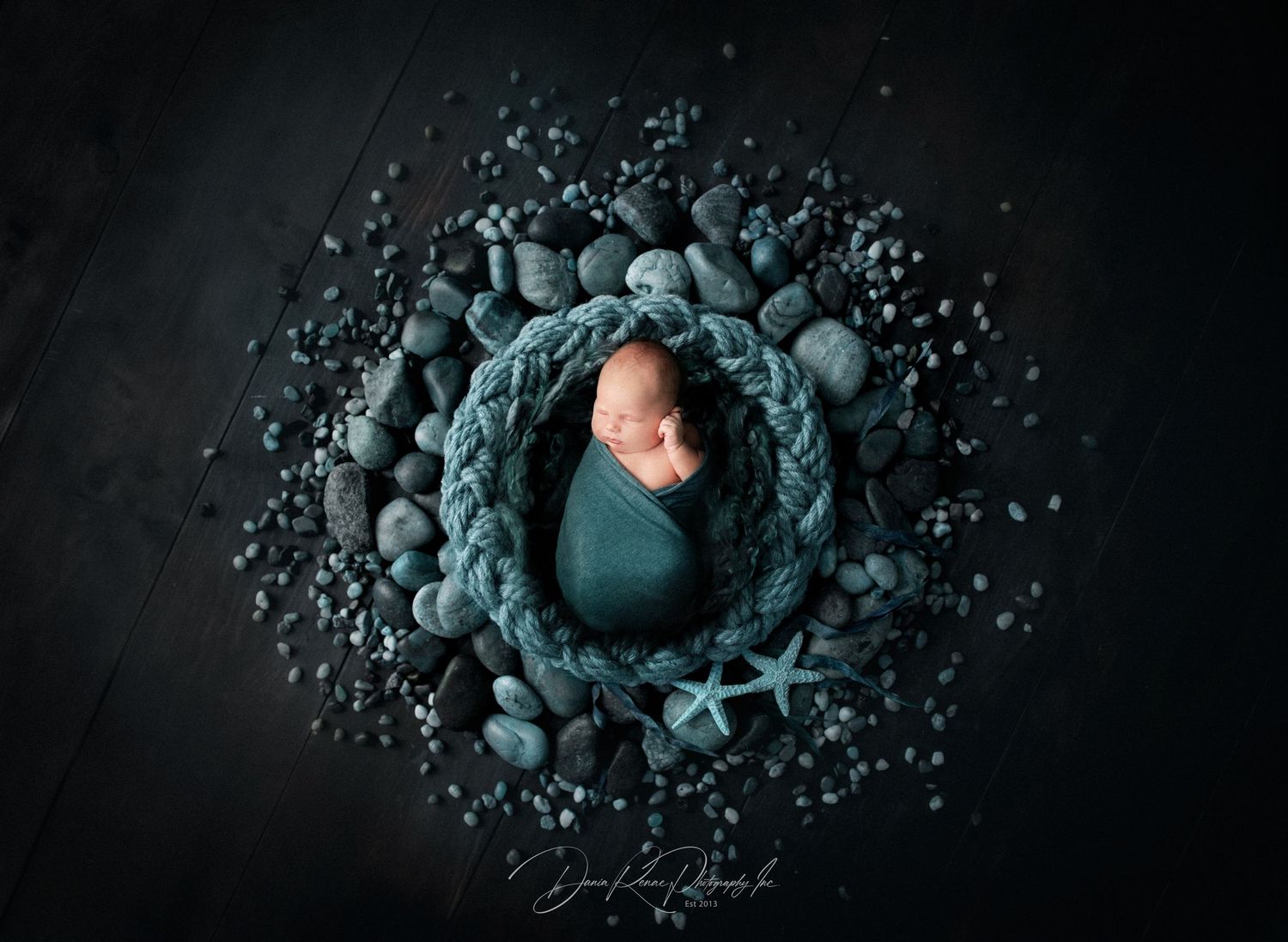Newborn baby swaddled in teal wrap, sleeping in braided rope bowl surrounded by gray pebbles and starfish.