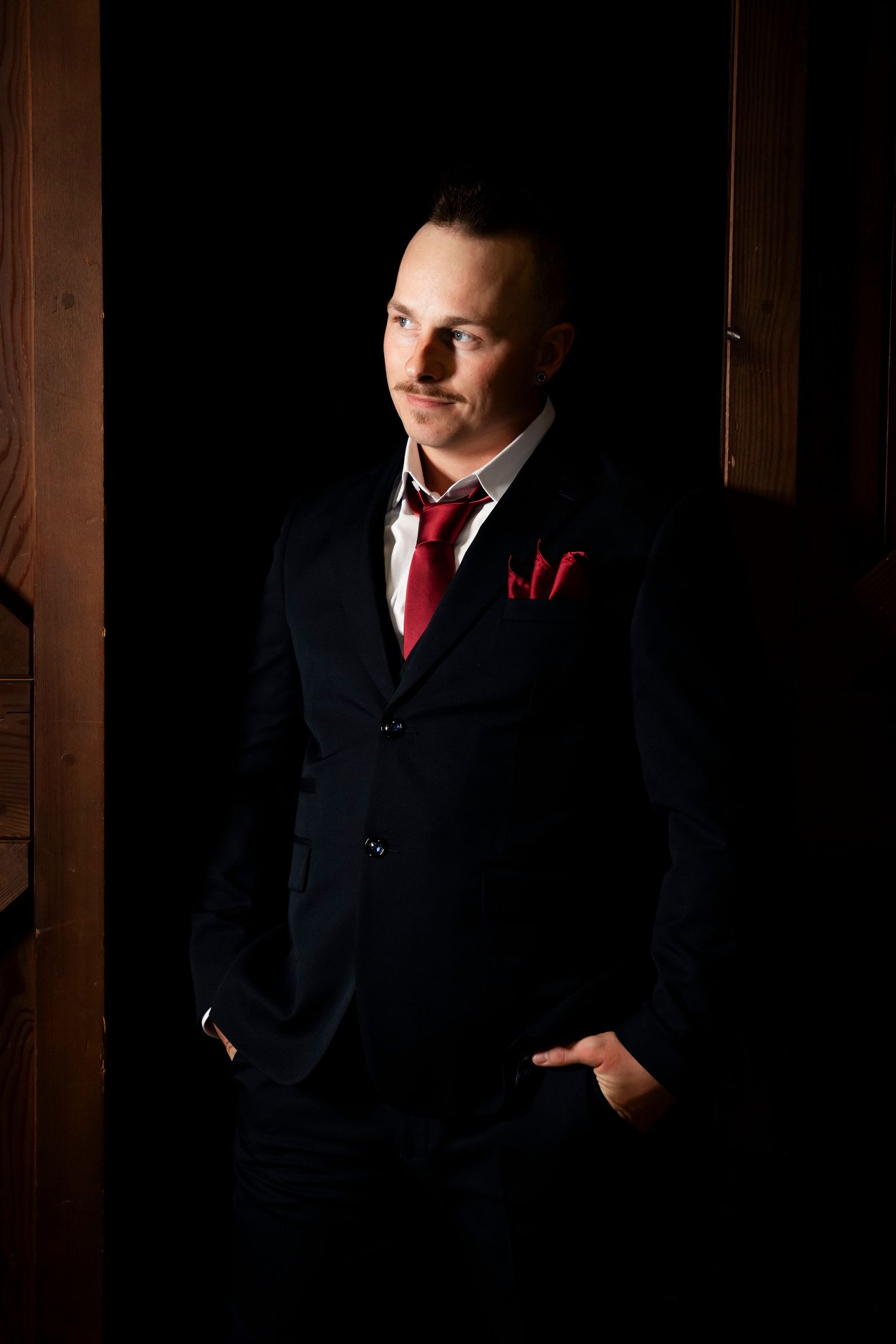 Elegant man in black suit with red tie and pocket square standing confidently in a dramatic dark doorway.