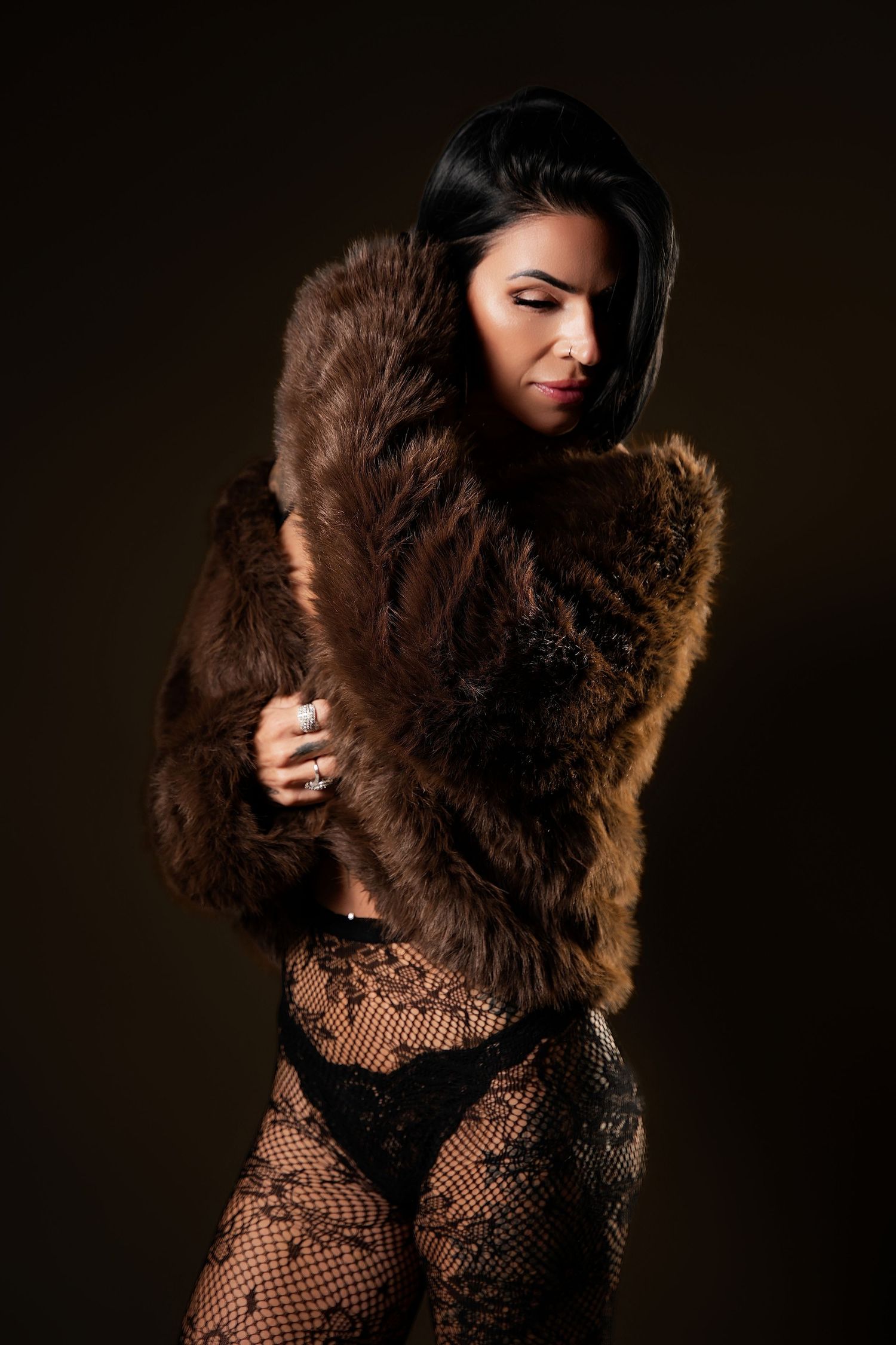 Woman in brown fur coat and black lace bodysuit posing against dark background.