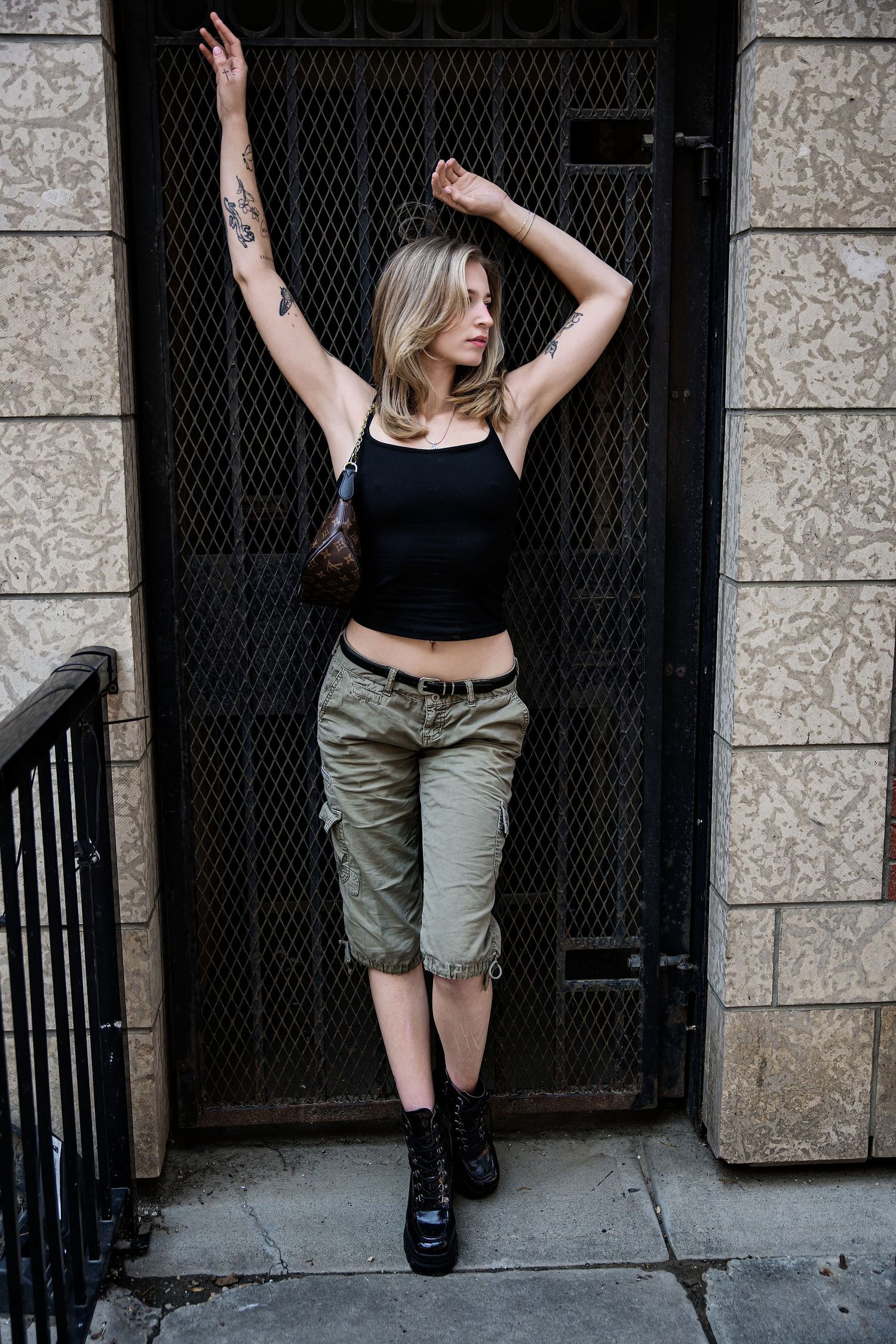 Young blonde woman with tattoos posing in black crop top, cargo capris, and boots against a metal gate.
