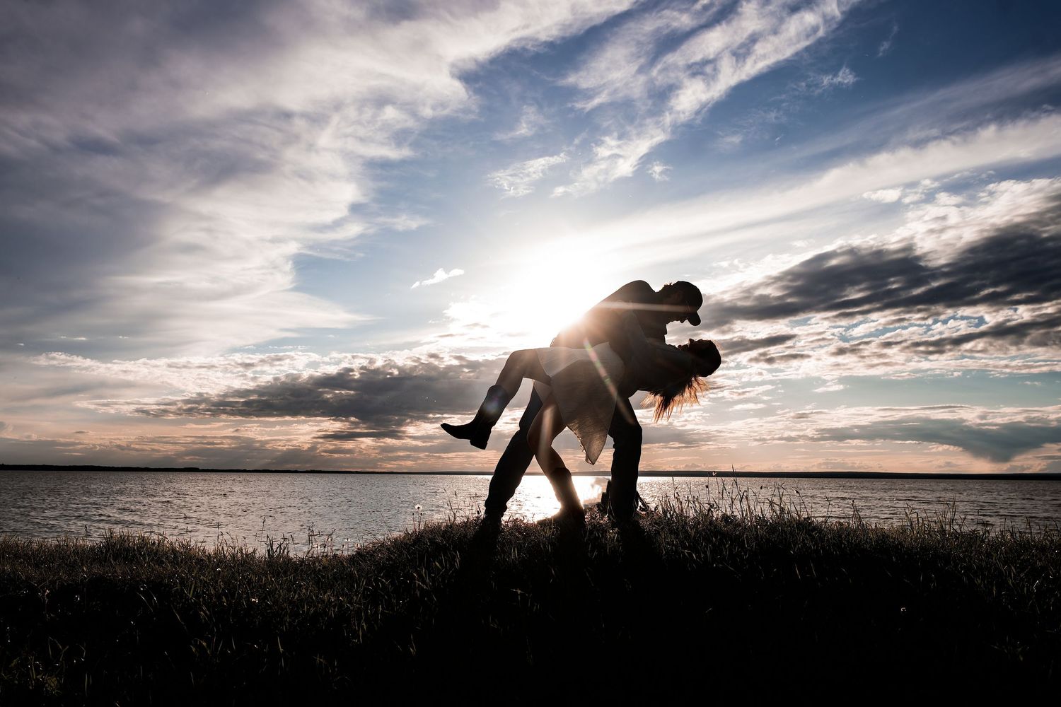 Silhouette of a couple dancing romantically by a lake at sunset with dramatic cloudy sky backdrop.