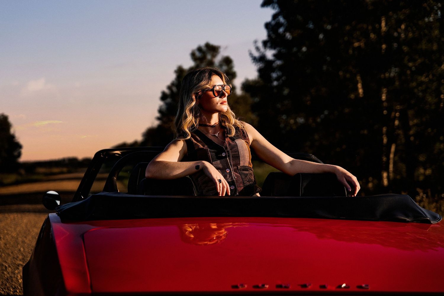Woman in sunglasses posing in a red convertible car at sunset on a rural road.