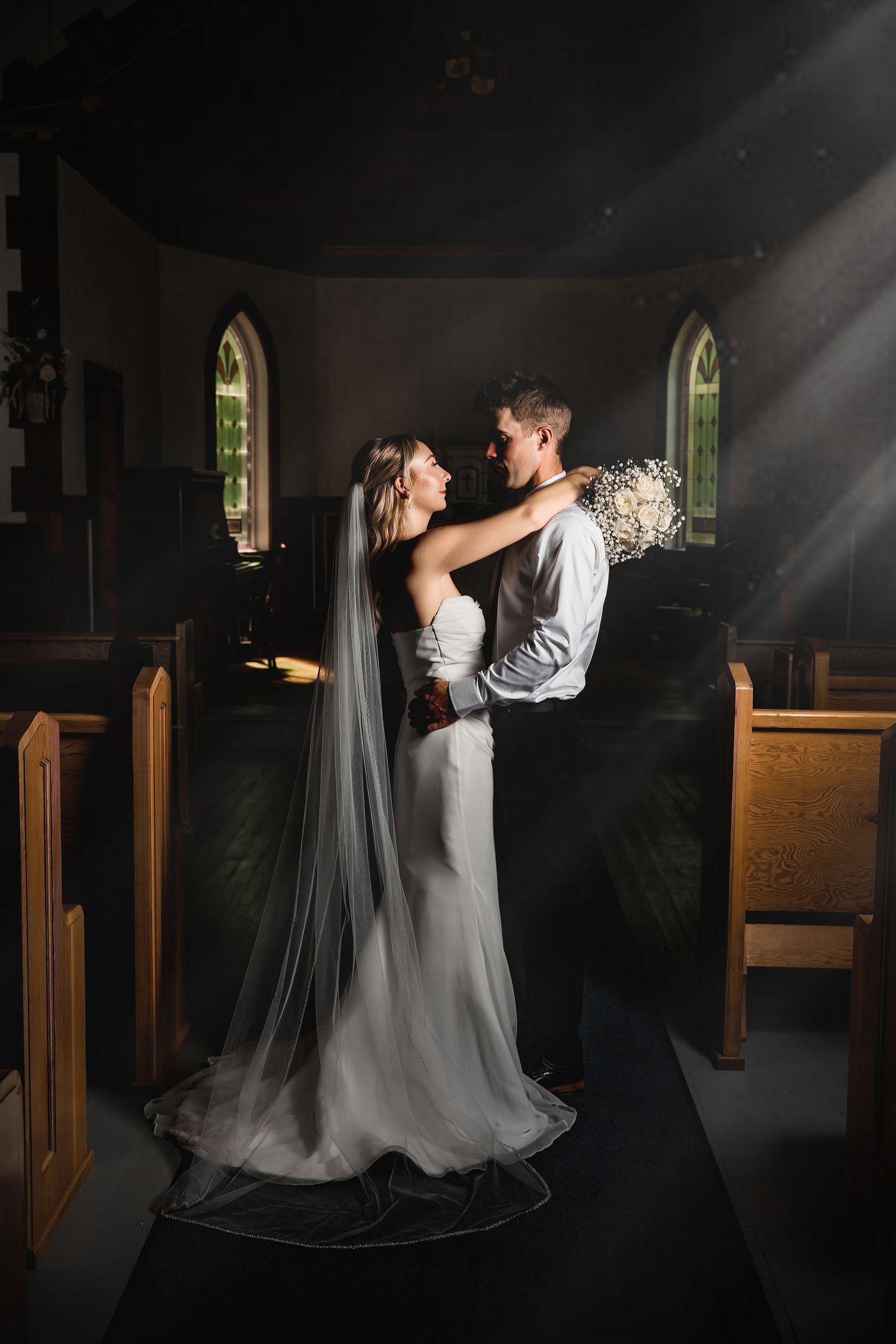 Bride and groom embrace in a sunlit church aisle, wedding veil trailing, holding a bouquet in a romantic ceremony.