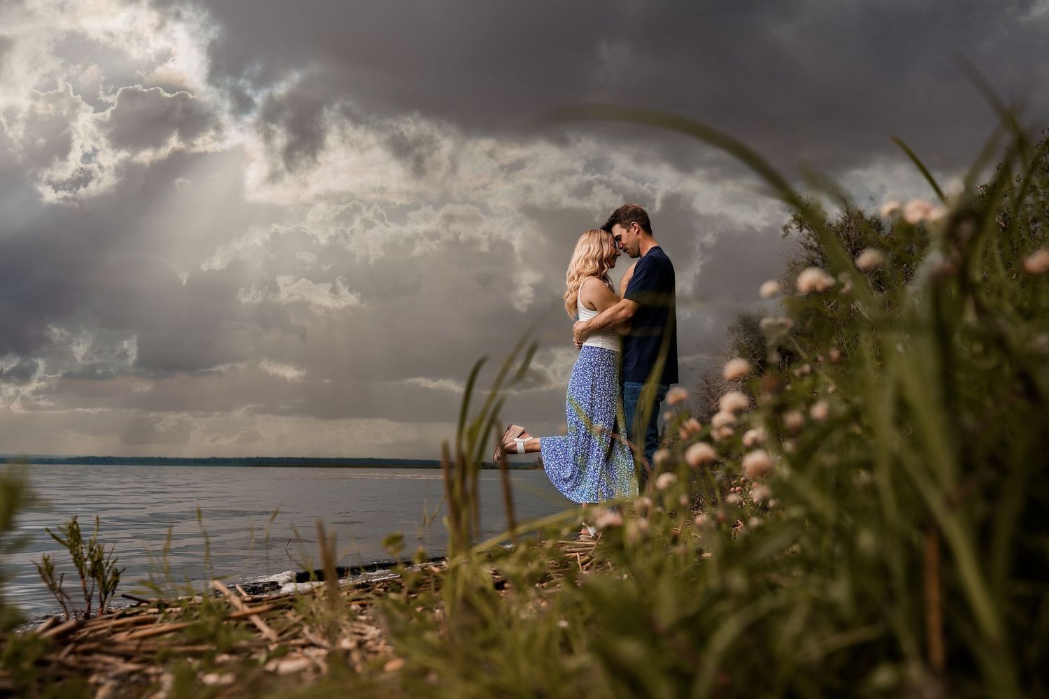 Couple embracing on a scenic waterfront cliff with dramatic stormy skies and wildflowers in the foreground.