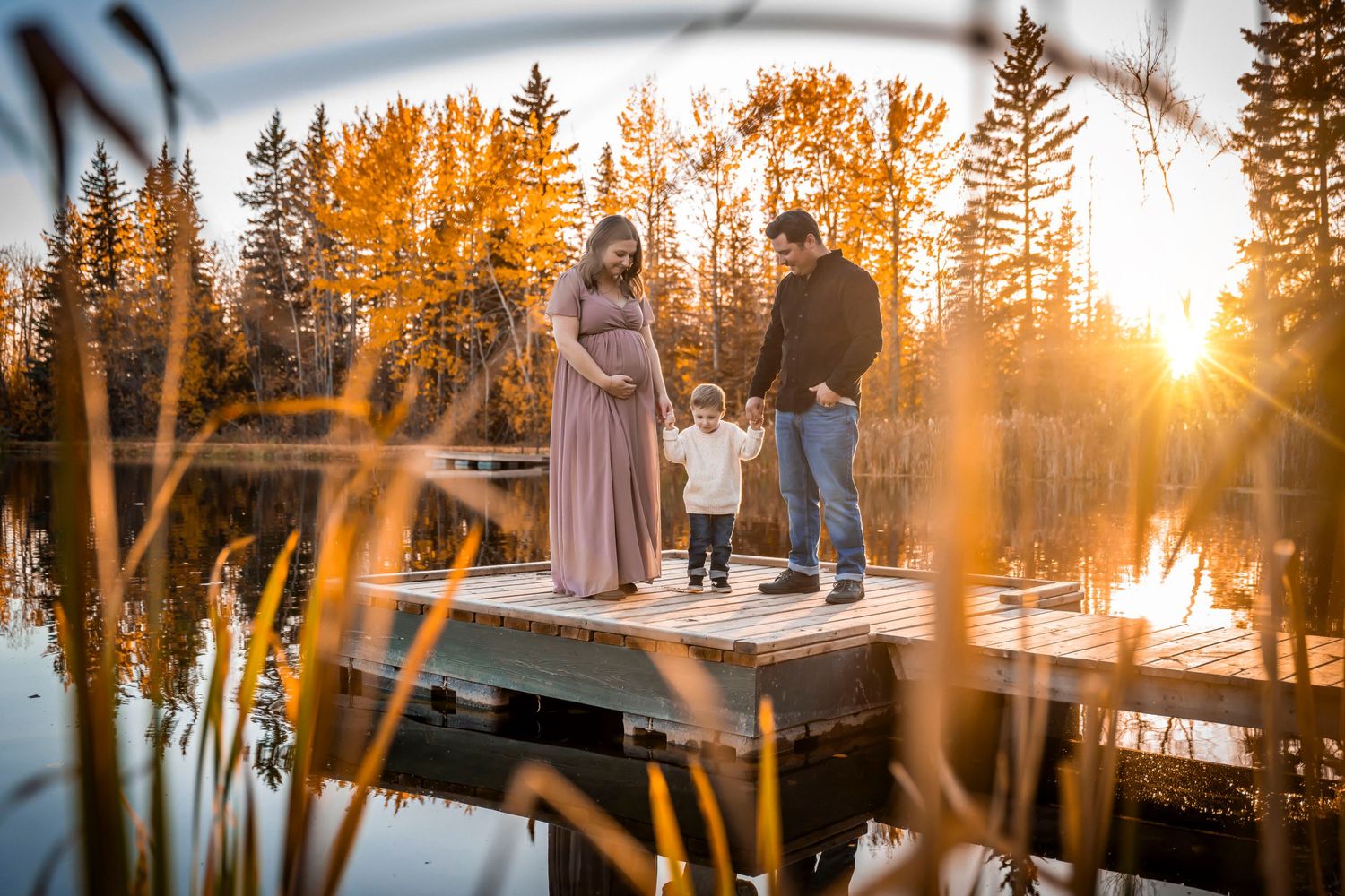 Pregnant woman in mauve gown with family on lake dock at golden sunset surrounded by autumn trees.