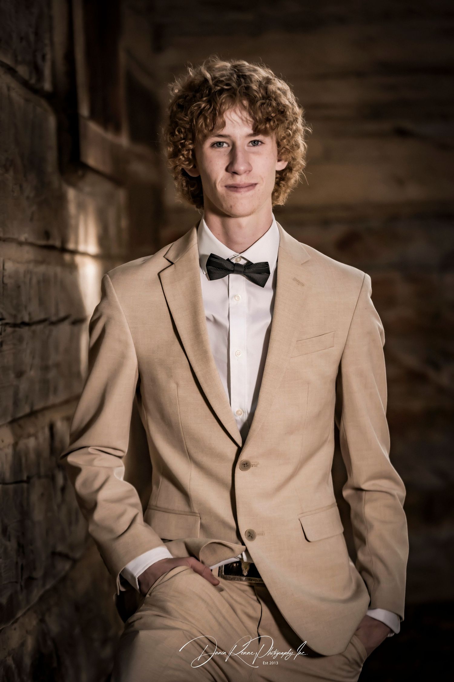 Young man in tan suit with black bow tie poses confidently against rustic stone wall in formal portrait.