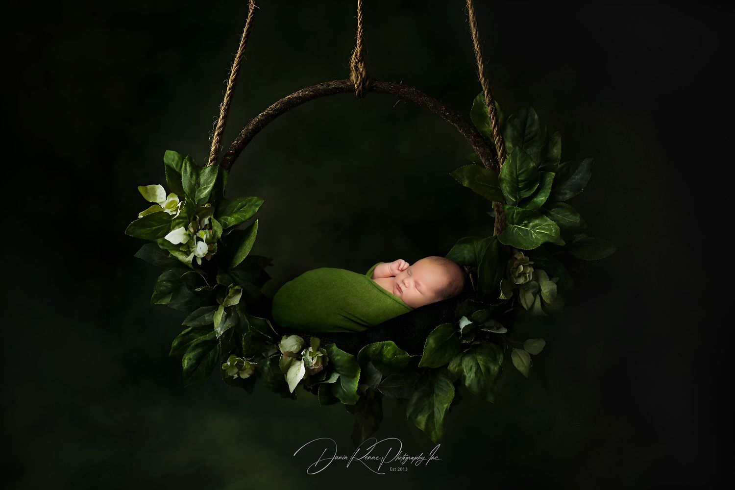 Newborn baby sleeping peacefully in a green wrap inside a floral wreath swing with white flowers on dark background.