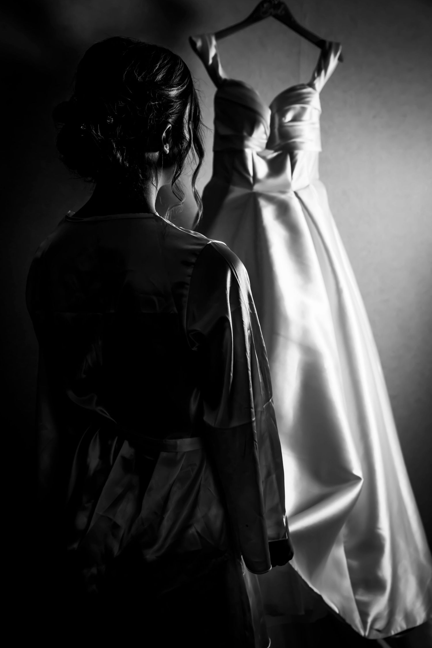 Black and white photo of a bride in a robe admiring her elegant strapless wedding gown hanging on the wall.