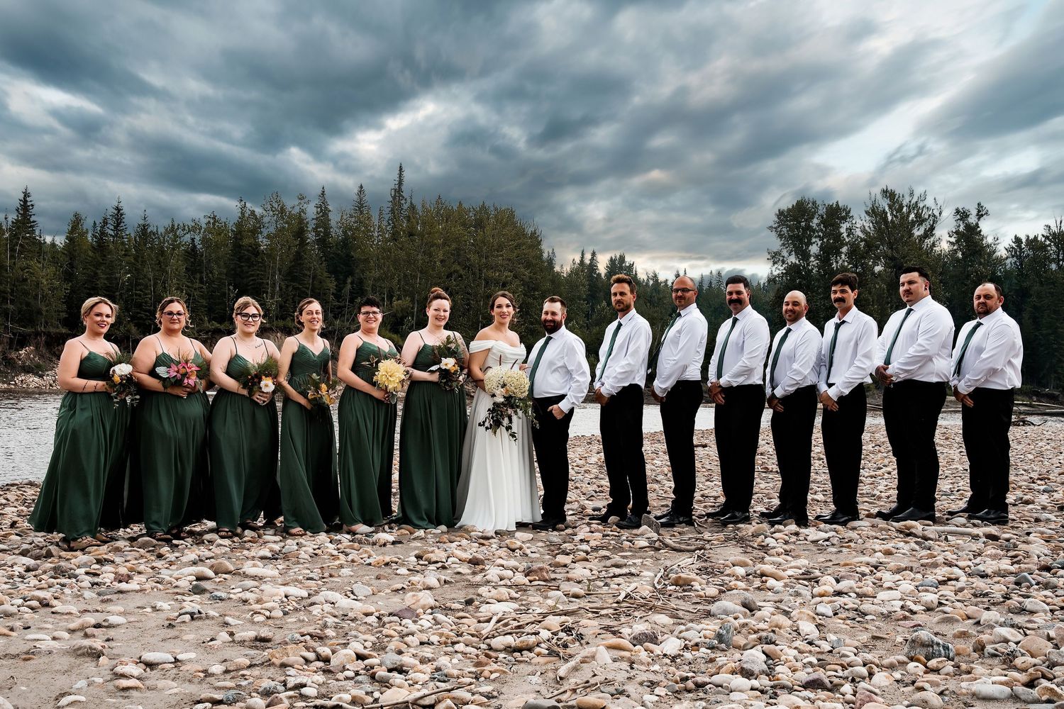 Wedding party poses on rocky riverbank with stormy sky, bridesmaids in green gowns and groomsmen in black suits.