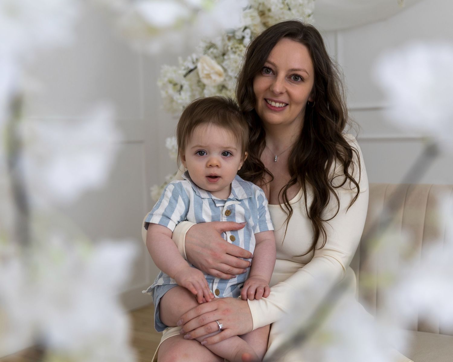 A smiling woman with long dark hair holds a baby boy in a blue outfit, surrounded by white floral decor.