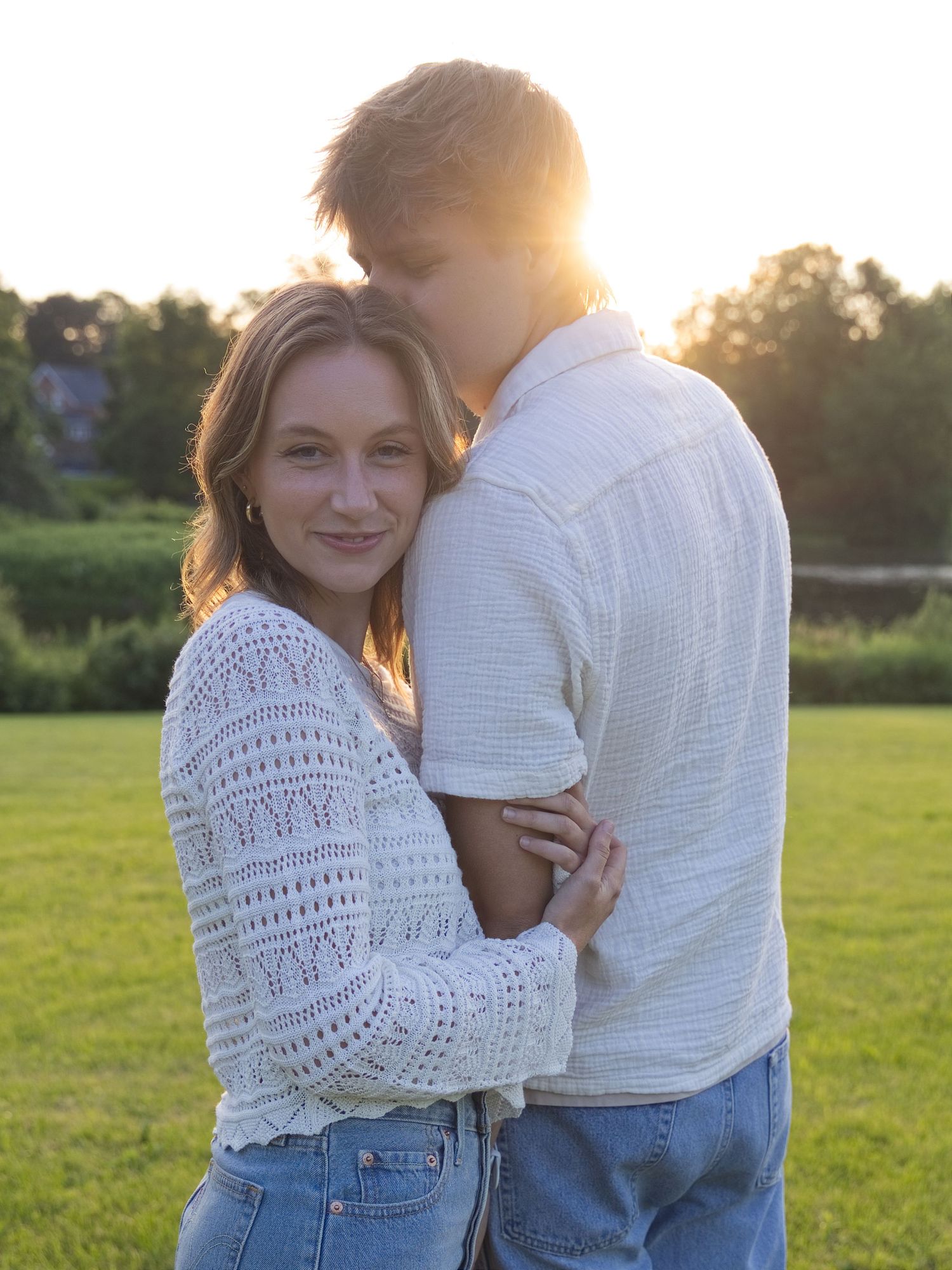 Couple embracing at golden hour sunset in a green field, woman in white lace top and jeans smiling warmly.