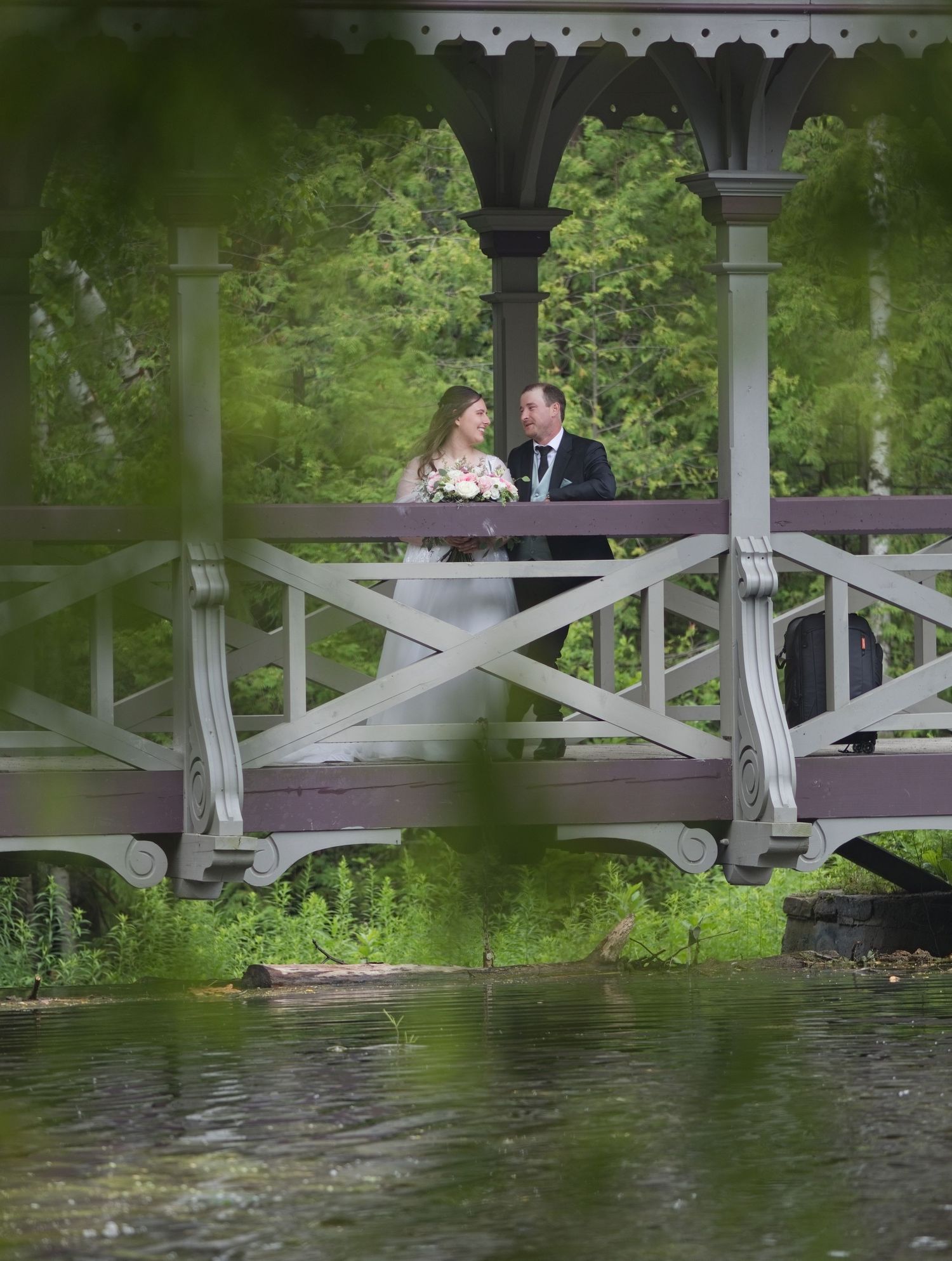 A bride and groom share a romantic moment on a white gazebo bridge surrounded by lush greenery and a tranquil pond.
