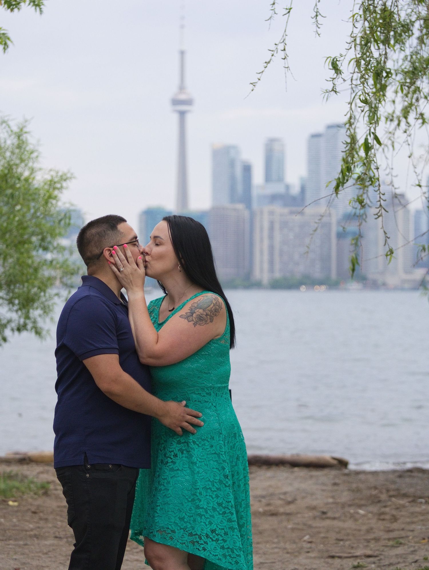 Couple kissing on Toronto waterfront with CN Tower and city skyline in background, woman in teal lace dress.