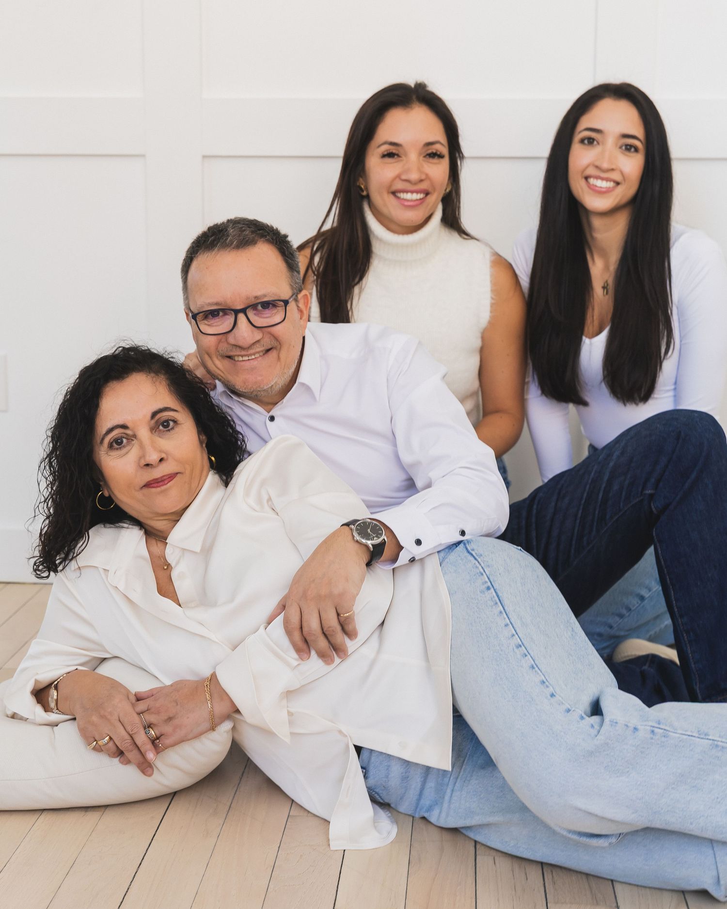 Family of four portrait with parents and two teenage daughters posing casually on a white floor in coordinated outfits.