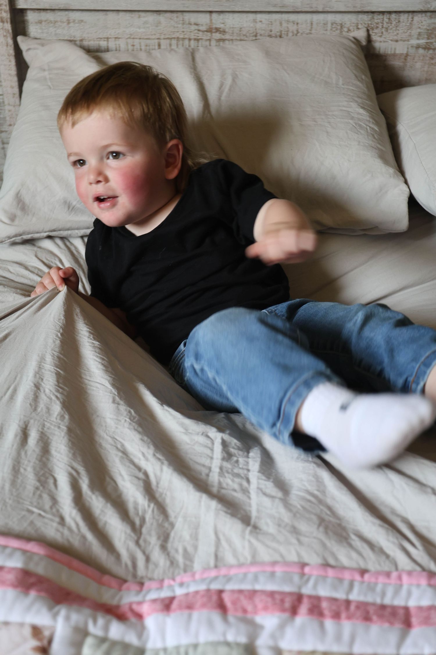 A red-cheeked toddler boy in a black shirt and jeans sits playfully on a bed with outstretched arm.