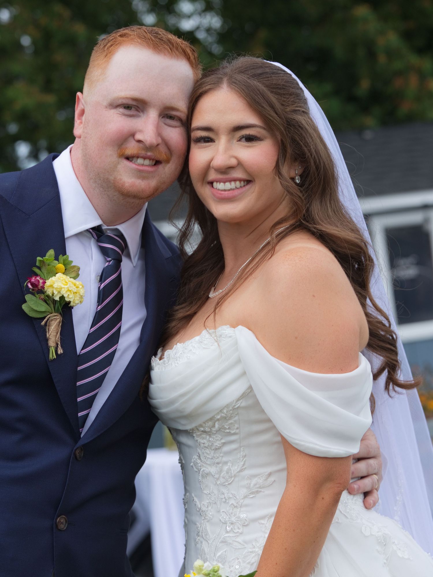 A smiling bride and groom pose together outdoors on their wedding day, she in a white off-shoulder gown and he in a navy suit.