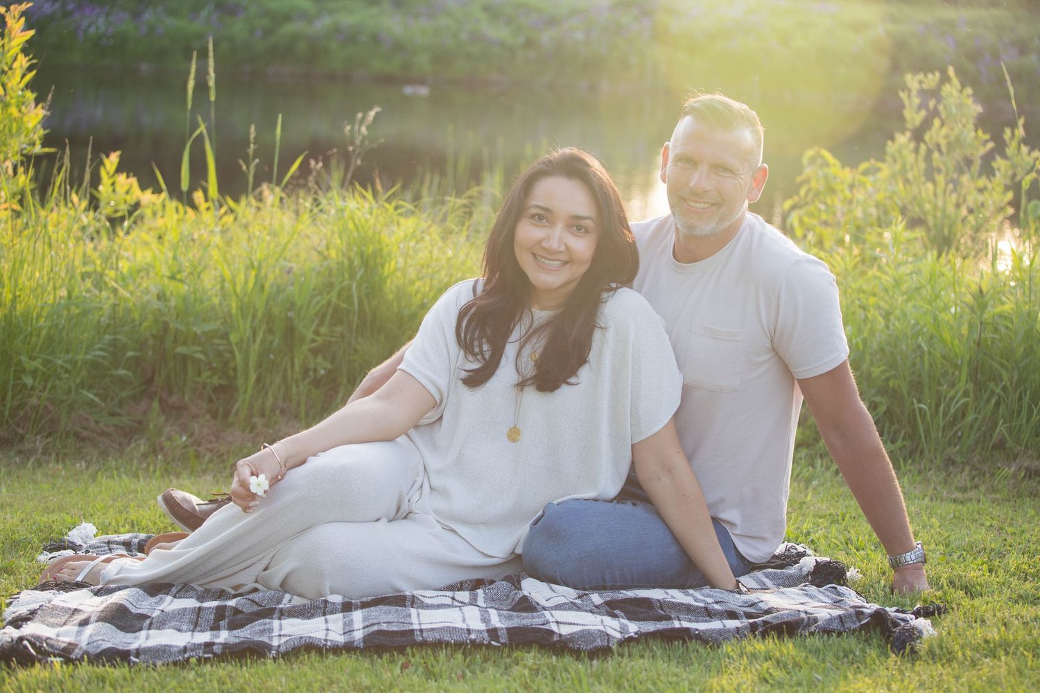 Couple sitting on plaid blanket in sunny green field, smiling warmly in casual white outfits during golden hour.