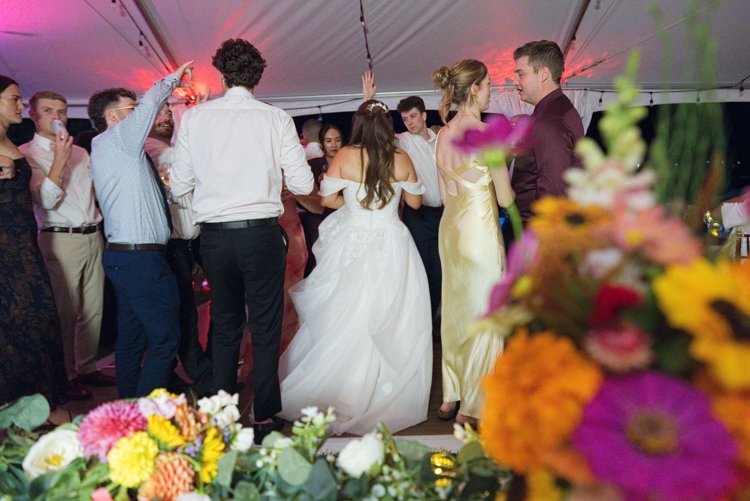 Wedding guests and bride dancing under tent at colorful floral reception celebration.