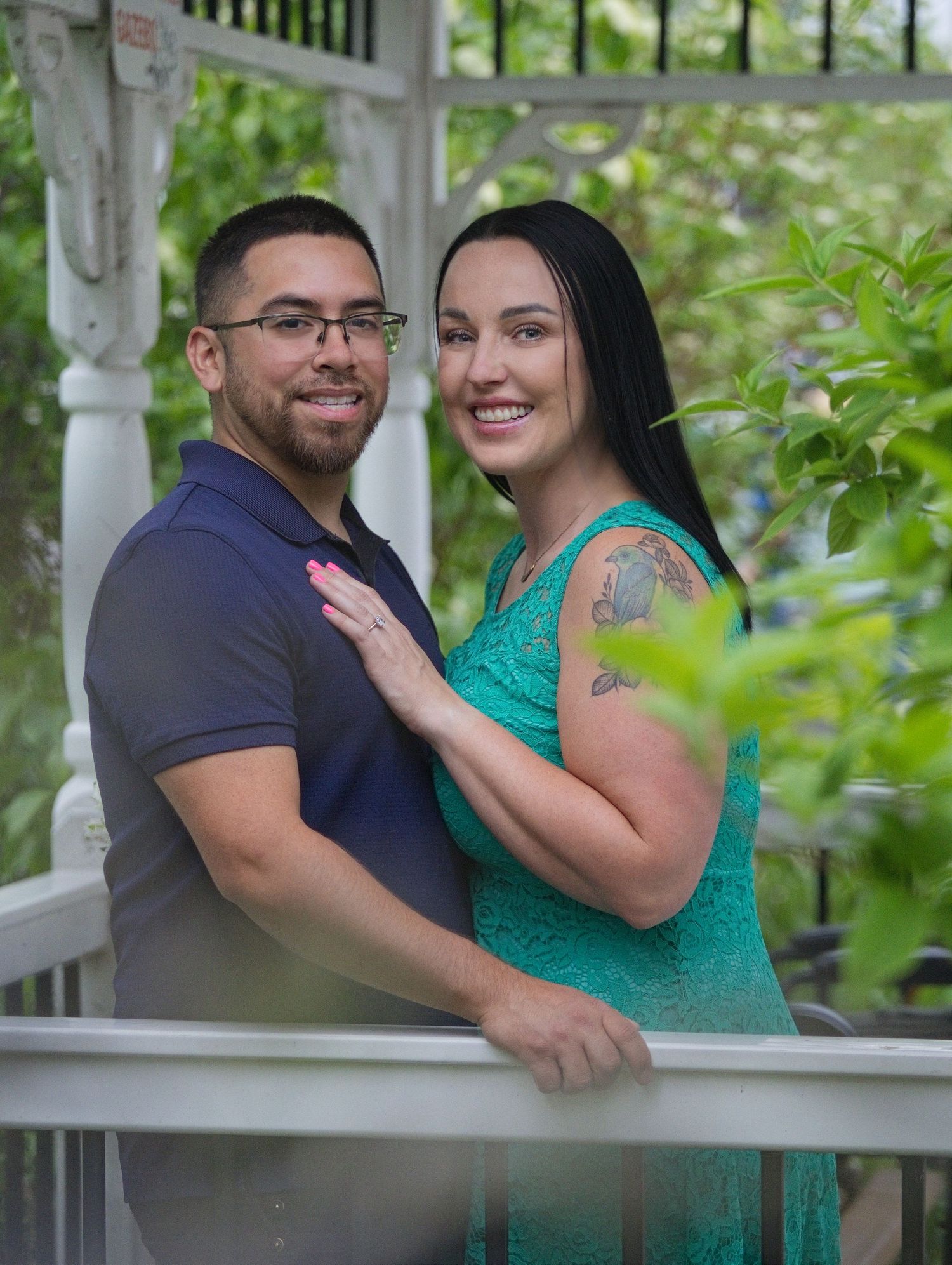 Smiling couple poses on a white gazebo porch surrounded by lush greenery, woman in teal dress with tattoo, man in navy polo.