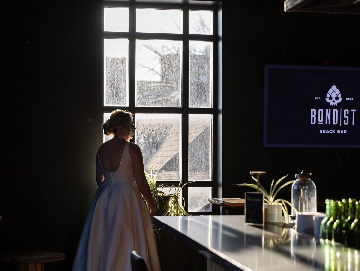 Bride in white gown standing at a bar near large windows at Bondist Snack Bar venue.