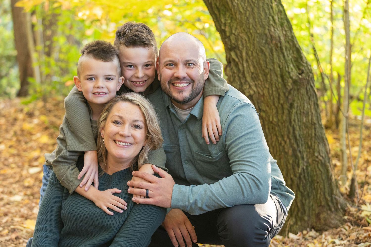 Family of four smiling outdoors in autumn forest, two boys hugging parents among golden fall foliage.
