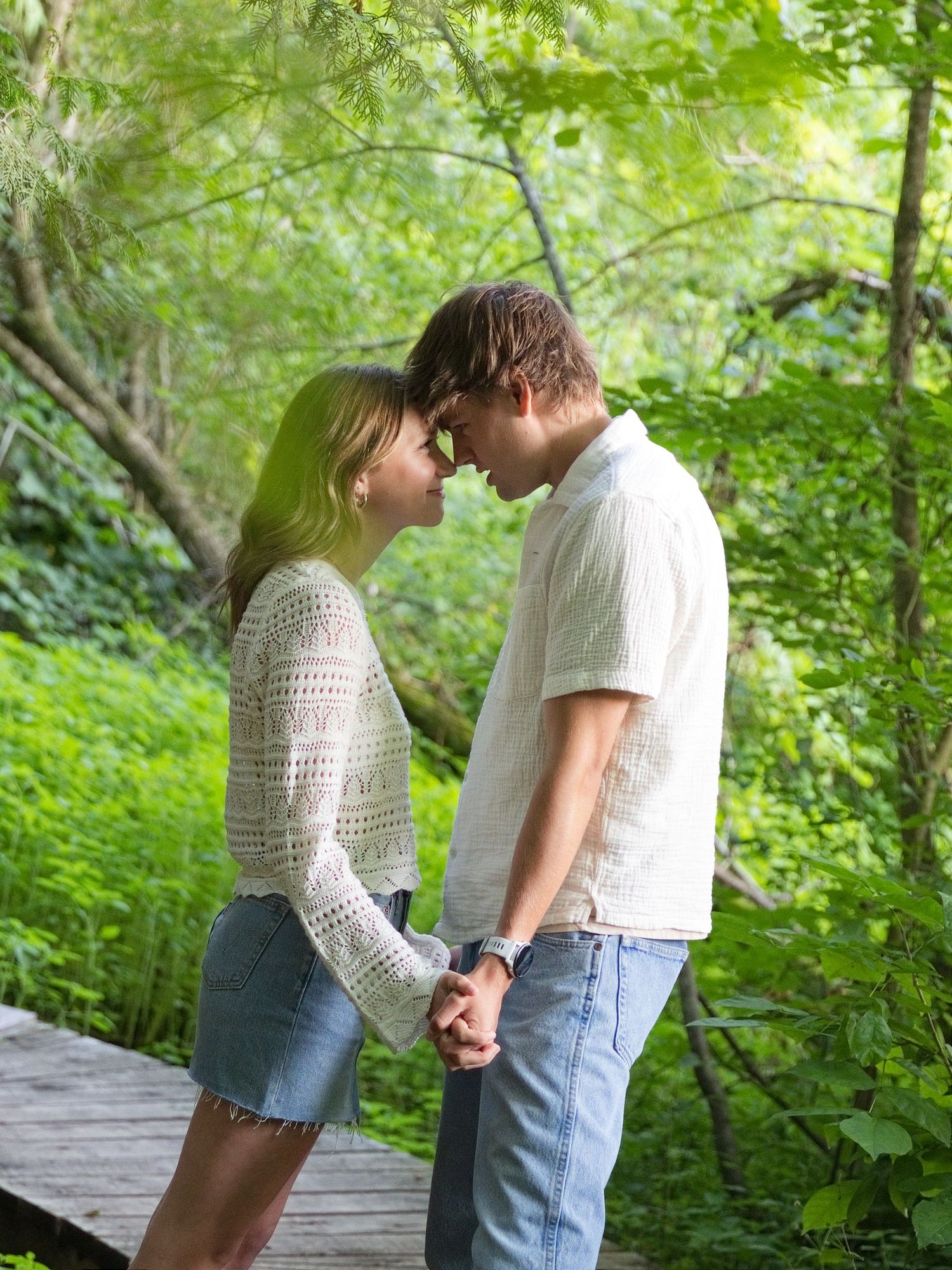 A young couple holds hands and leans in for a kiss on a wooden bridge surrounded by lush green trees.