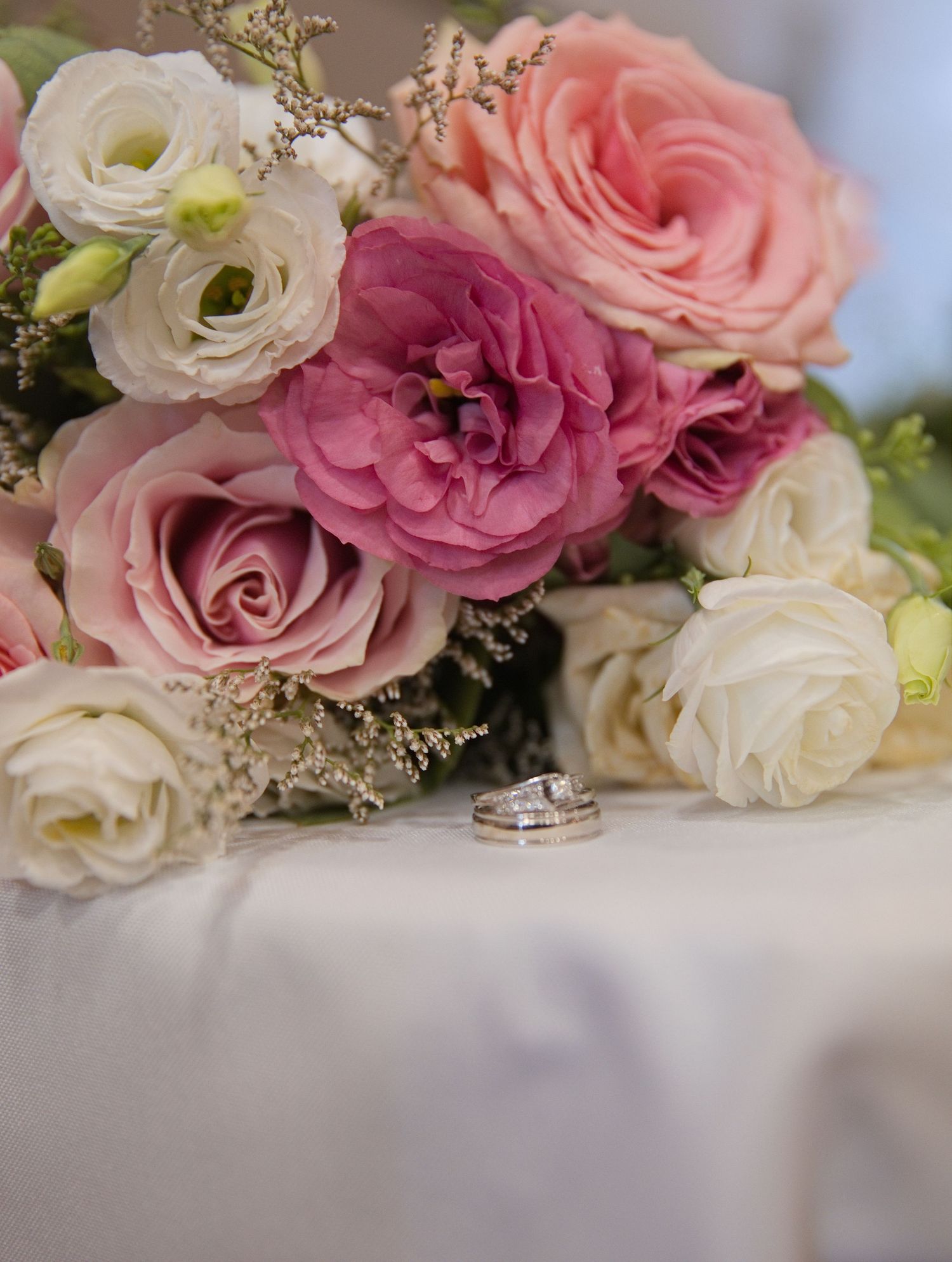 Wedding rings rest beside a romantic bouquet of pink, mauve, and white roses on a white tablecloth.