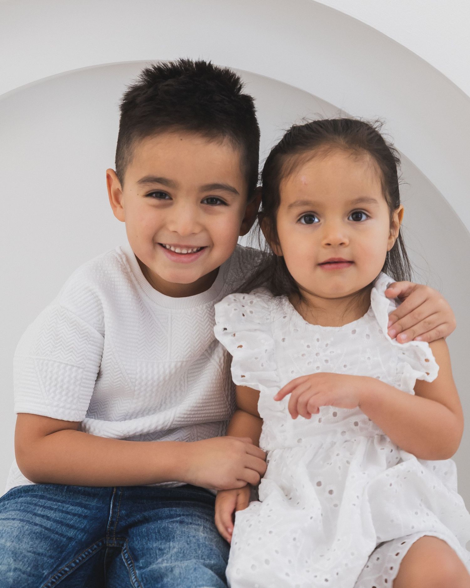A smiling young boy hugs his little sister in a white eyelet dress during a studio portrait session.