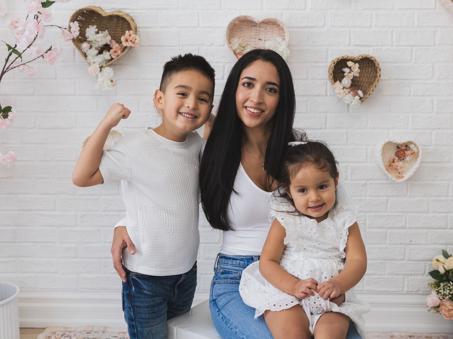 Mother posing with her young son flexing his arm and toddler daughter against a white wall with heart decorations.