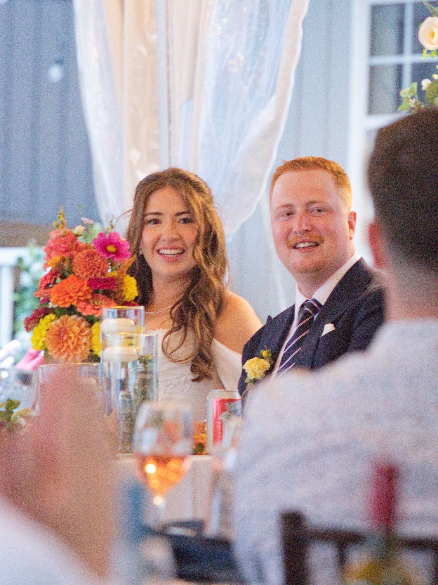Bride and groom smiling at wedding reception table with colorful floral centerpiece.