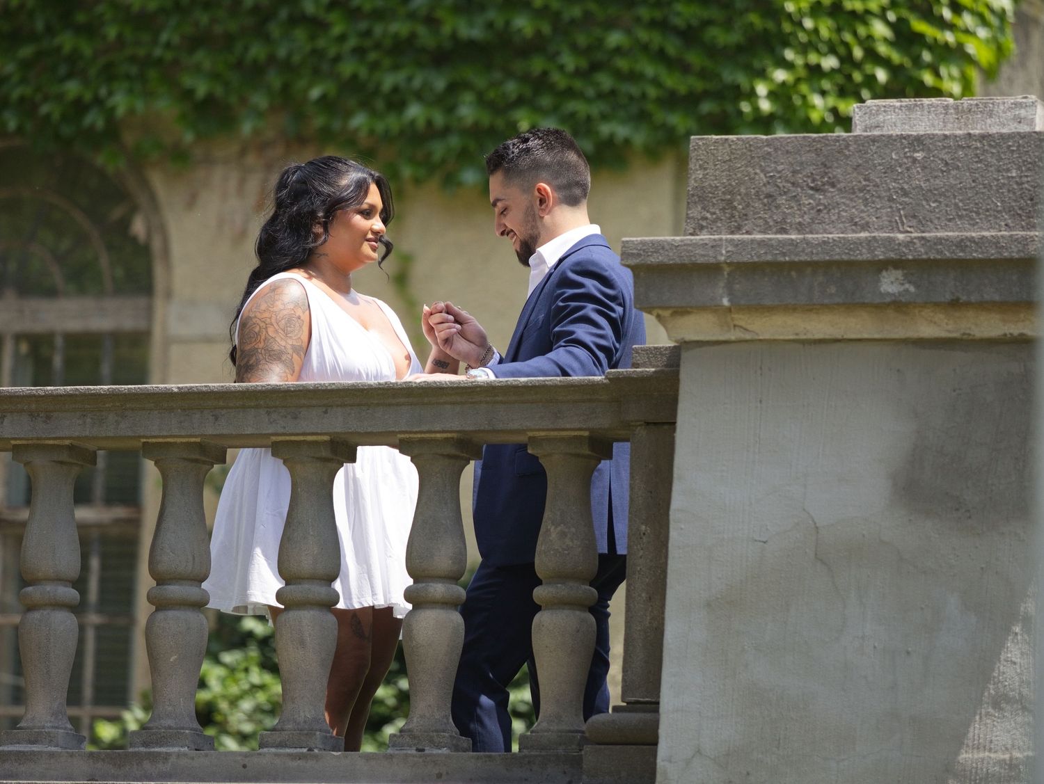 Couple in wedding attire holding hands on a stone balcony surrounded by lush greenery.