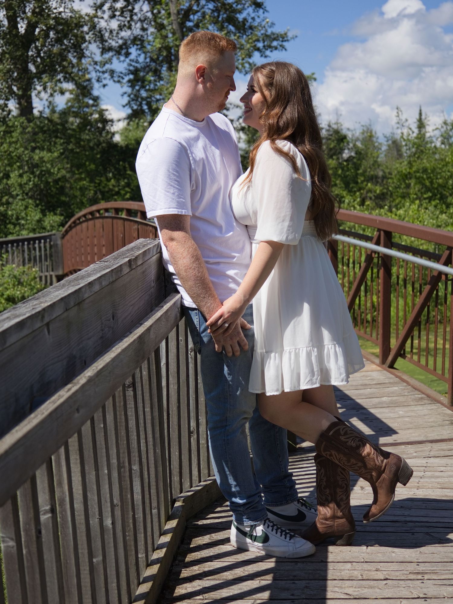 A couple in white outfits holds hands on a wooden bridge surrounded by lush green trees on a sunny day.