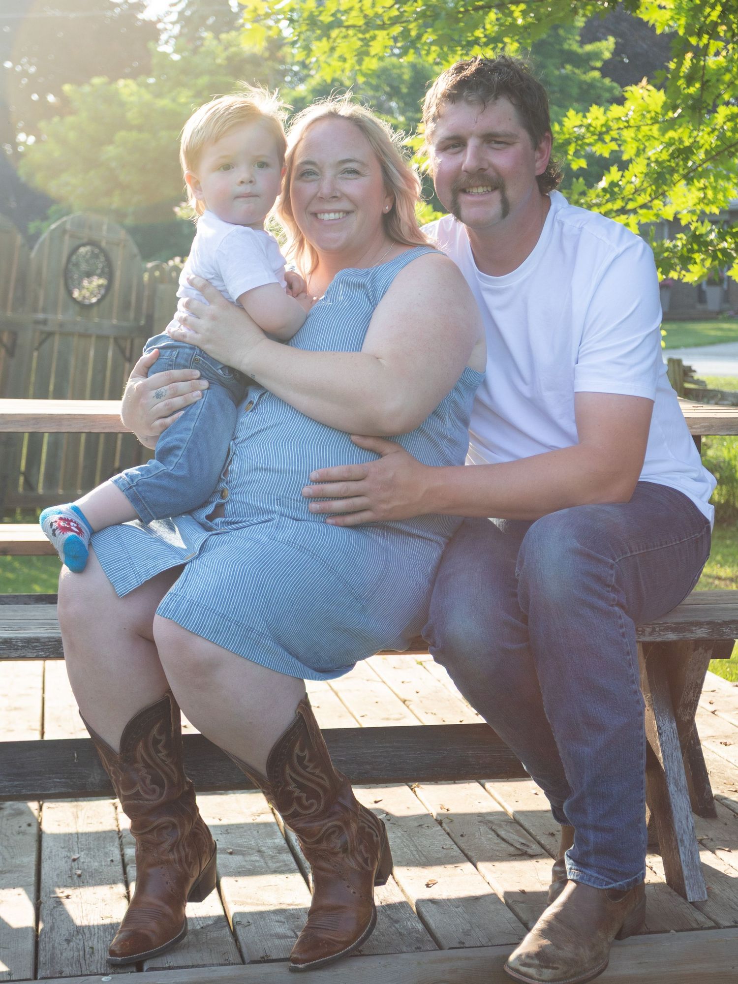 A smiling family of three poses on a wooden deck, with a woman in a blue dress and cowboy boots holding a toddler.