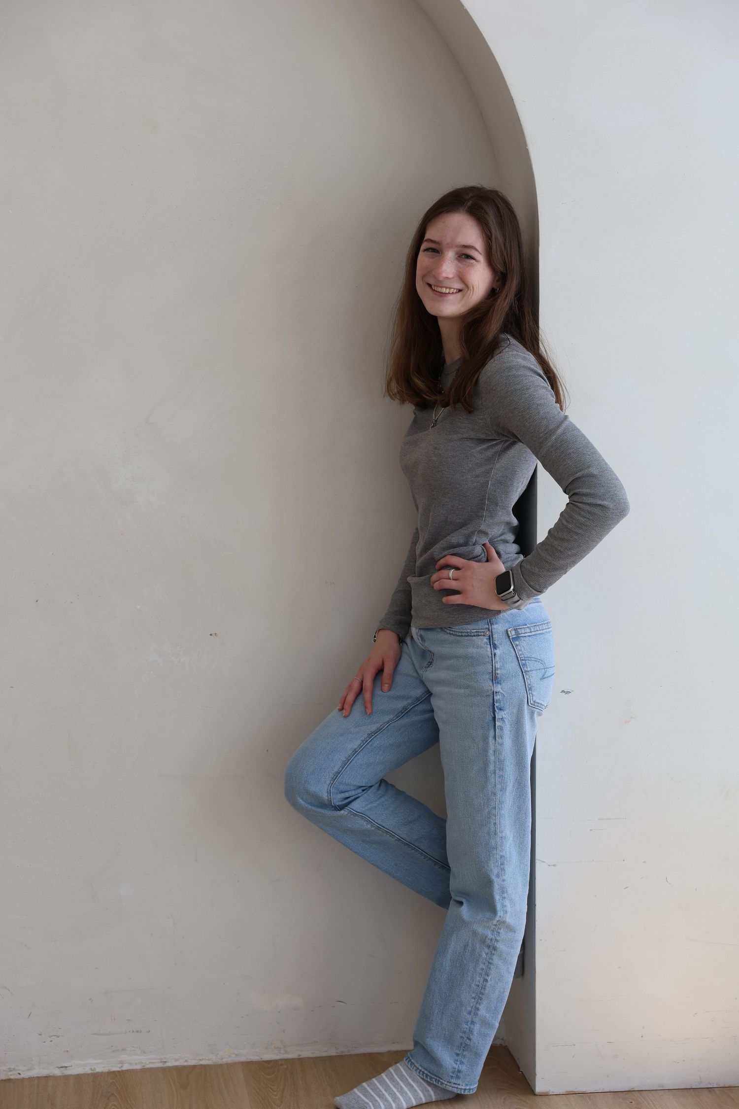 Smiling teenage girl in gray long-sleeve top and light blue jeans posing against a white arched wall.