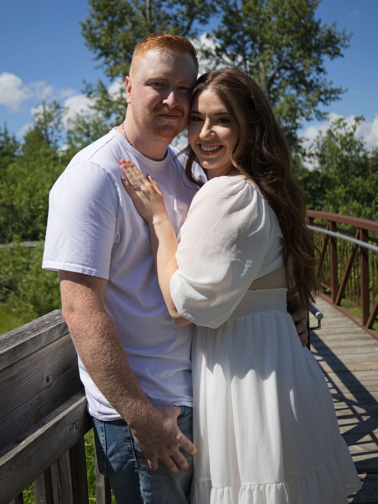 A smiling couple poses on a wooden bridge outdoors, woman in white dress, man in white tee, surrounded by lush greenery.