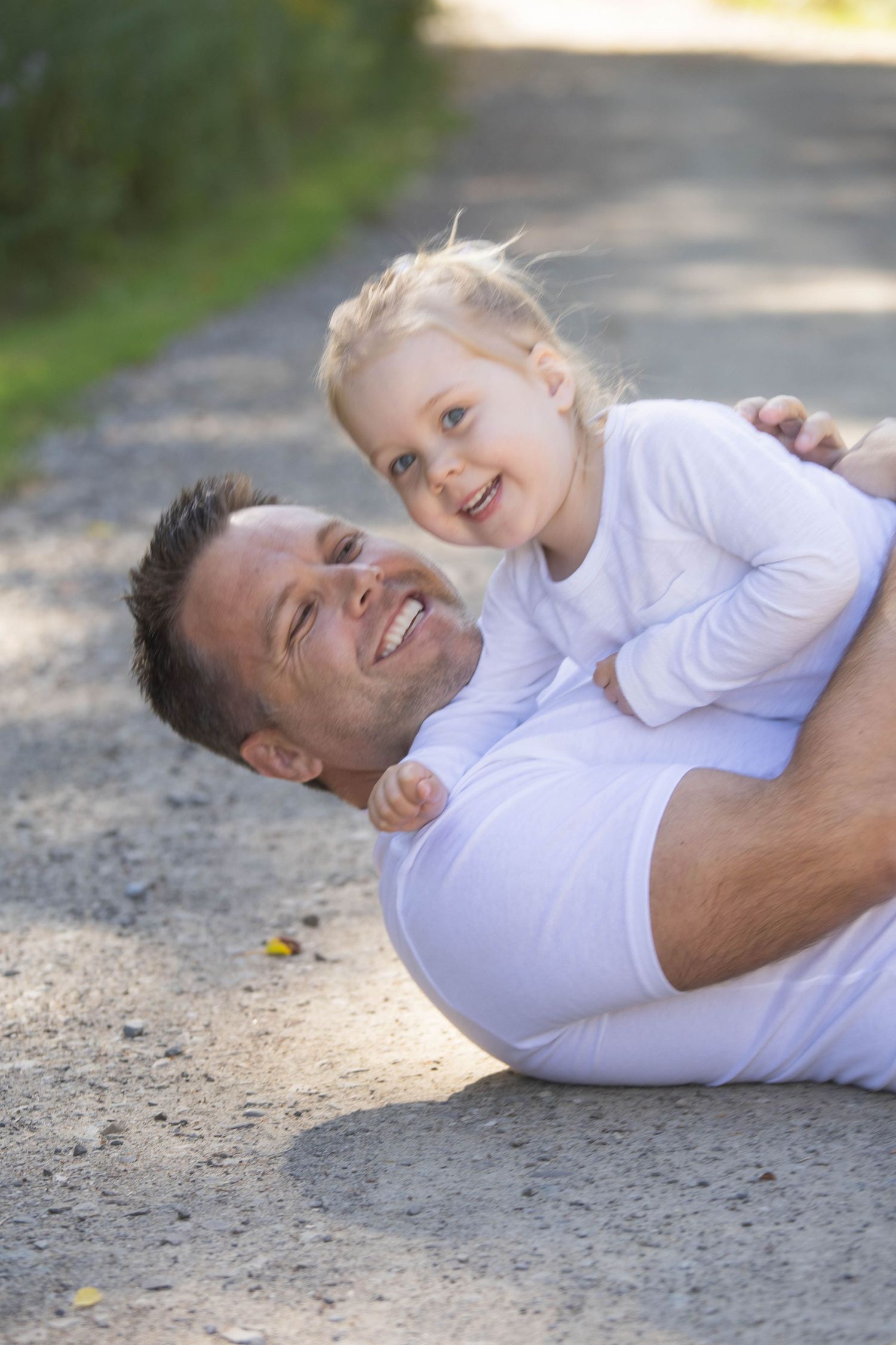 Joyful father lying on a path playfully holding his laughing blonde toddler outdoors.