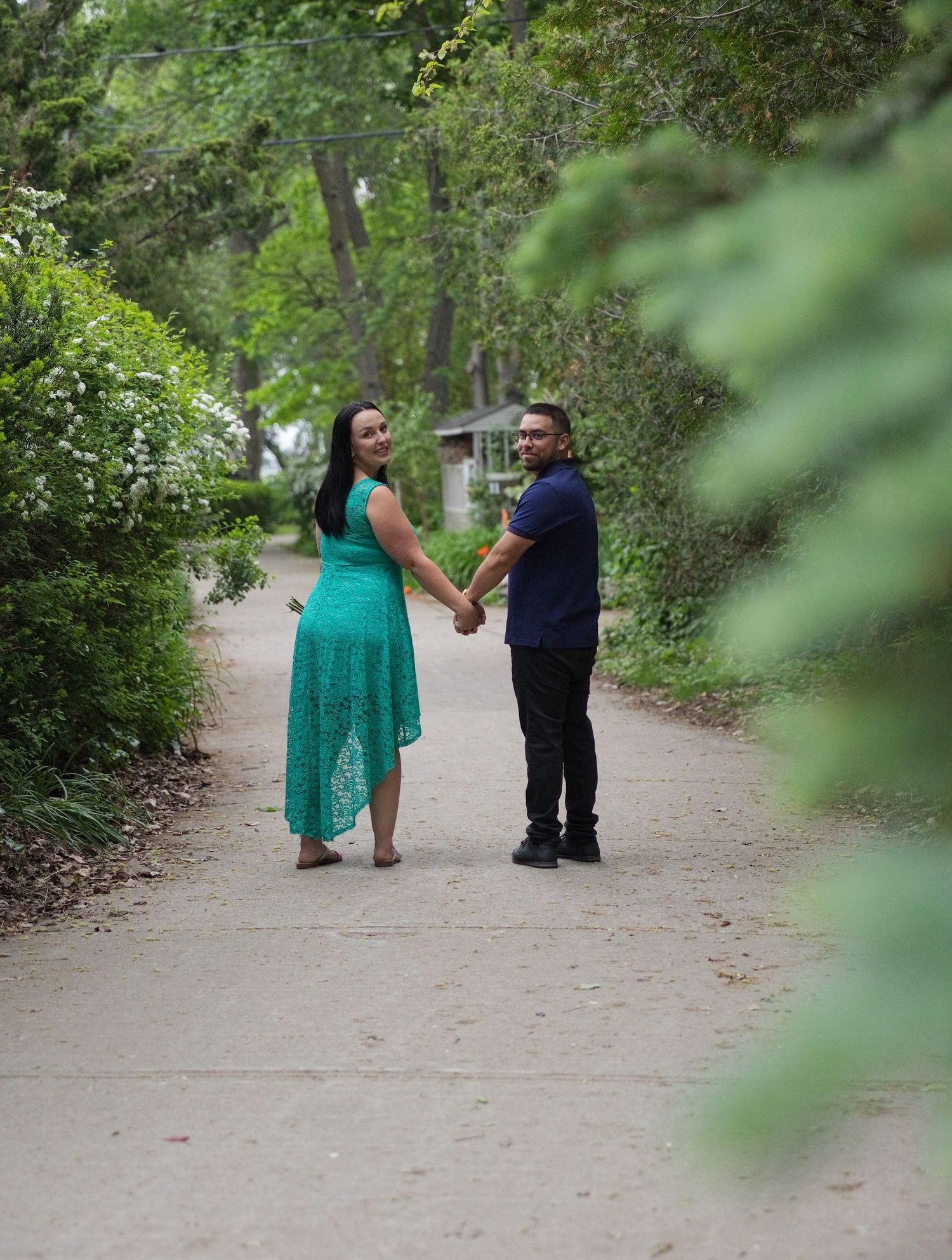Couple holding hands on a scenic tree-lined path, woman in teal dress and man in navy shirt, looking back at camera.