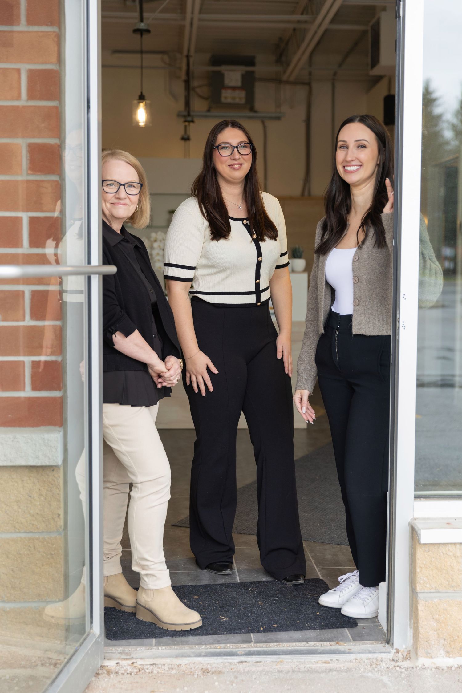 Three professional women smiling and posing together in a modern office doorway, dressed in smart business casual attire.