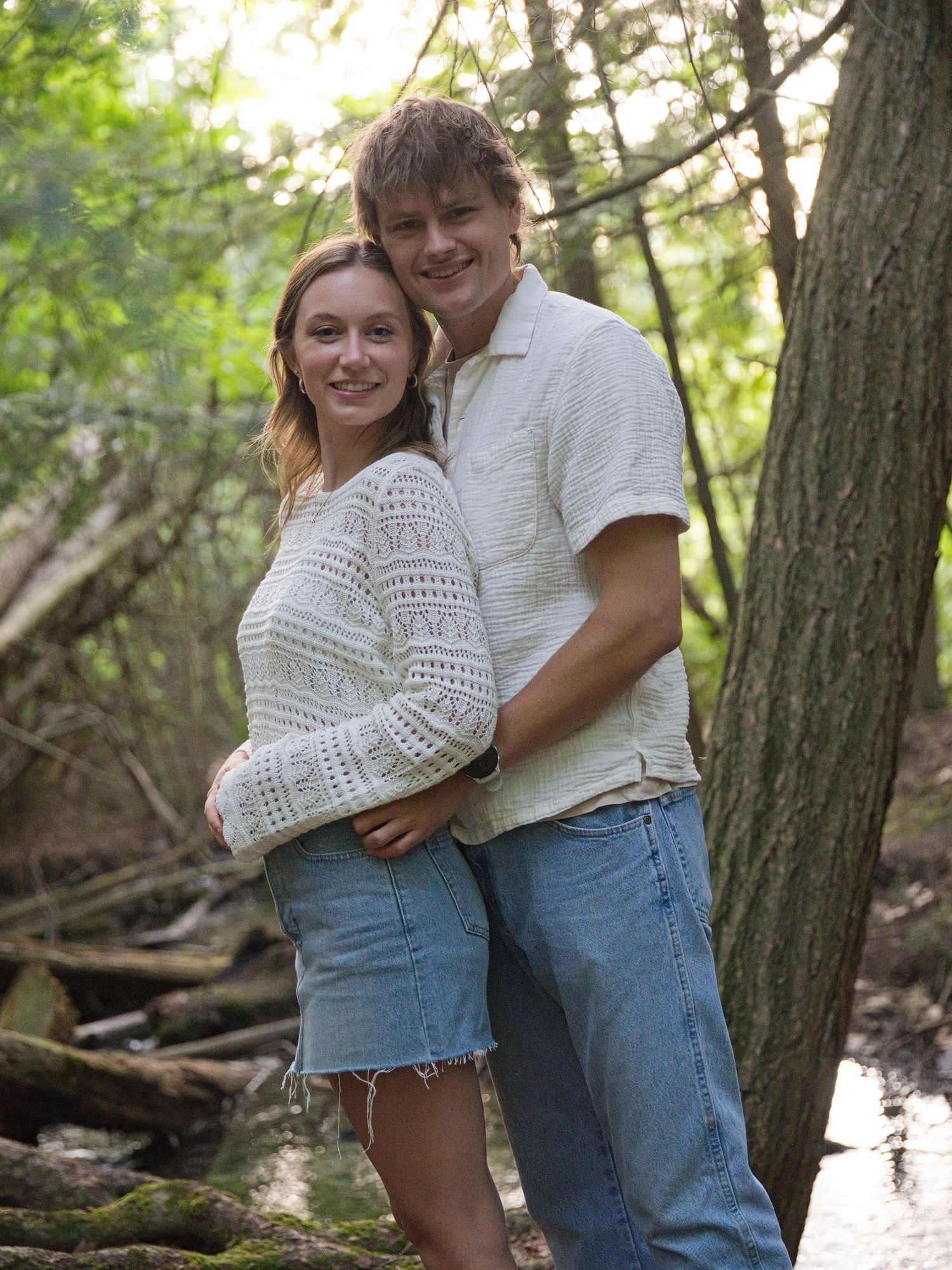 A young couple smiles together in a wooded area, both wearing white tops and jeans near a small creek.