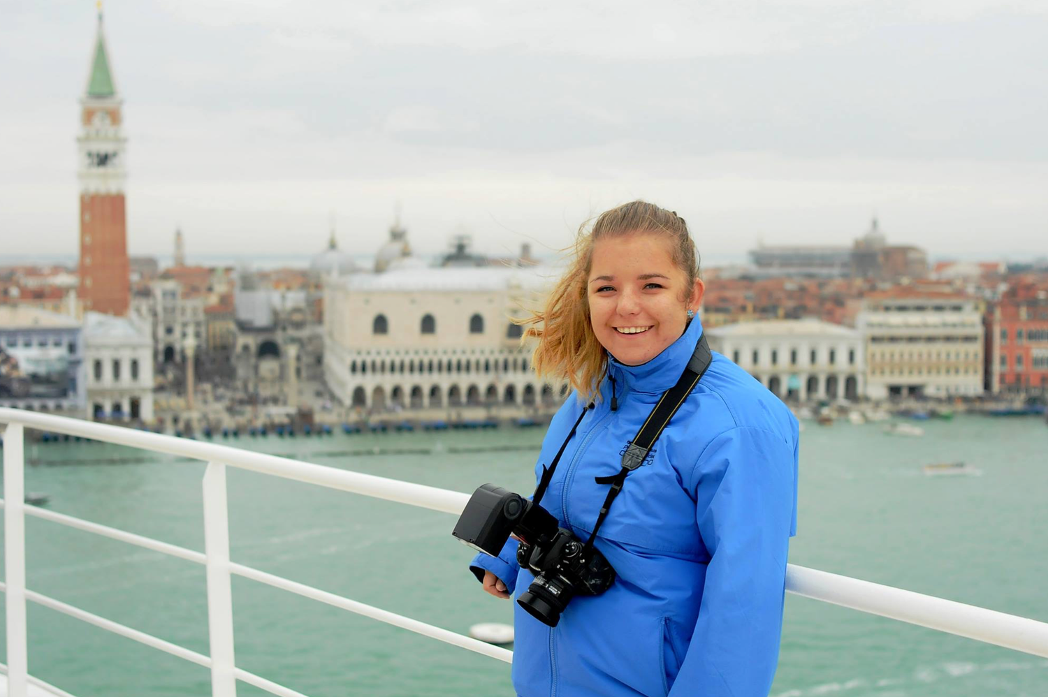 A smiling young woman with a camera stands on a boat deck with Venice's iconic skyline behind her.